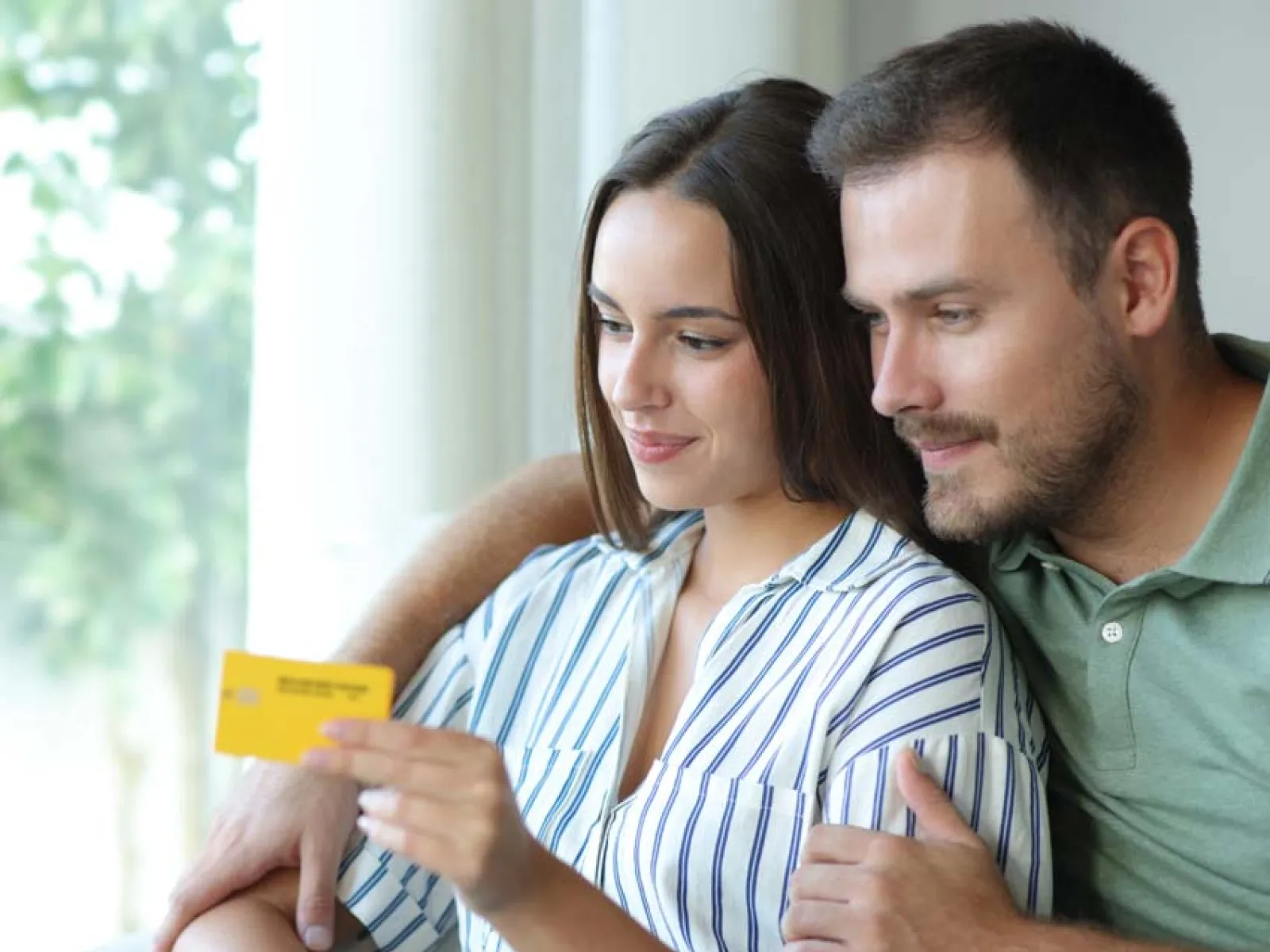 Couple shopping online using a laptop and credit card sitting together on a couch in a bright room
