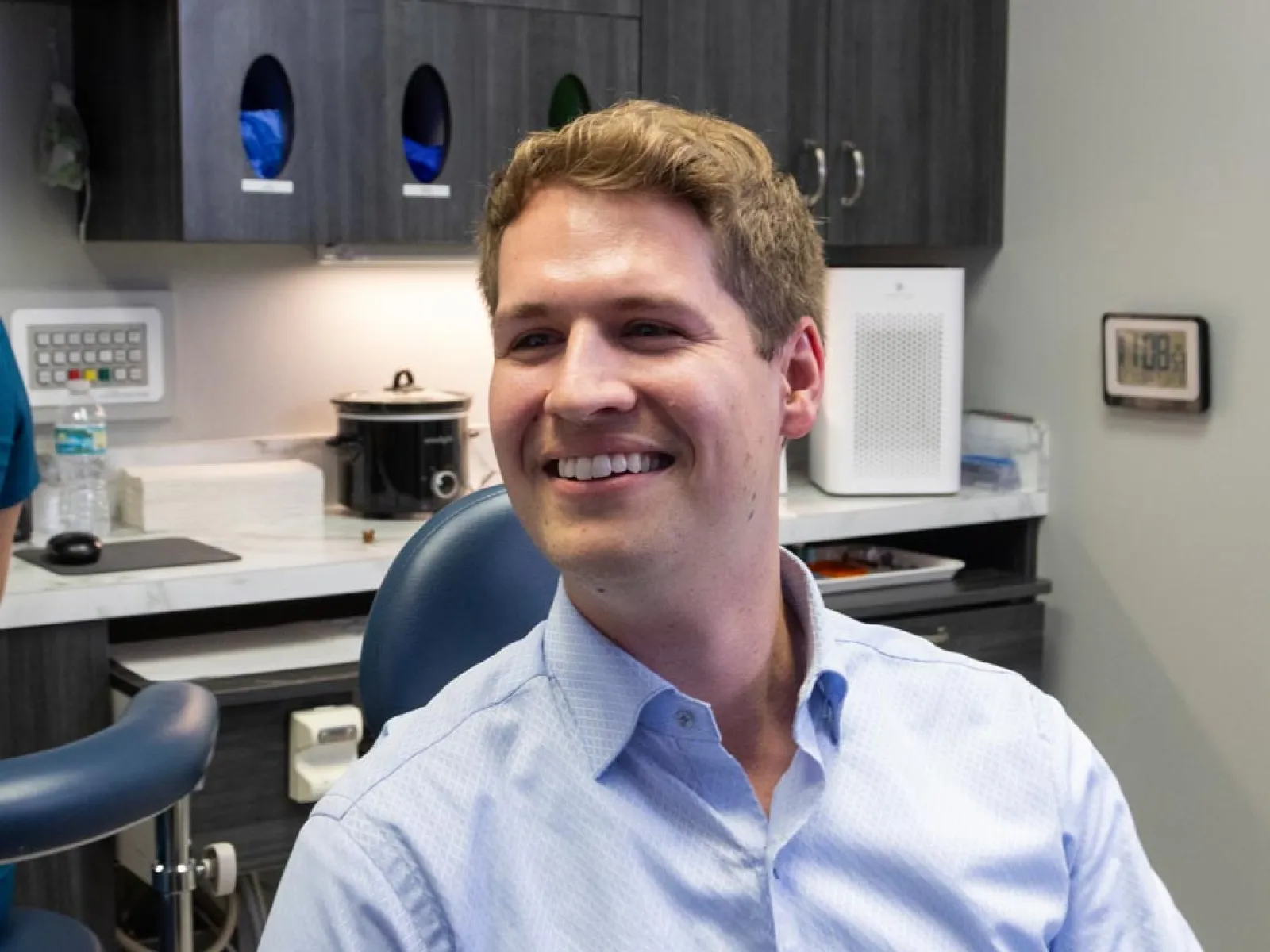 Smiling man in light blue shirt sitting in a modern dental clinic with cabinets and equipment behind him
