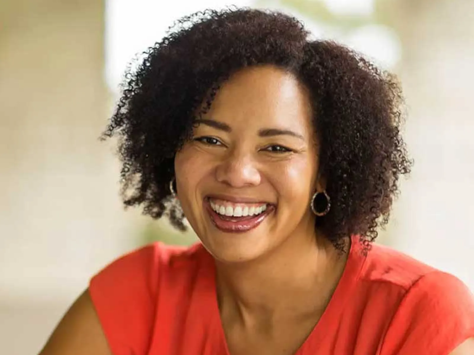 Smiling woman with curly hair wearing a red blouse and hoop earrings in a bright indoor setting