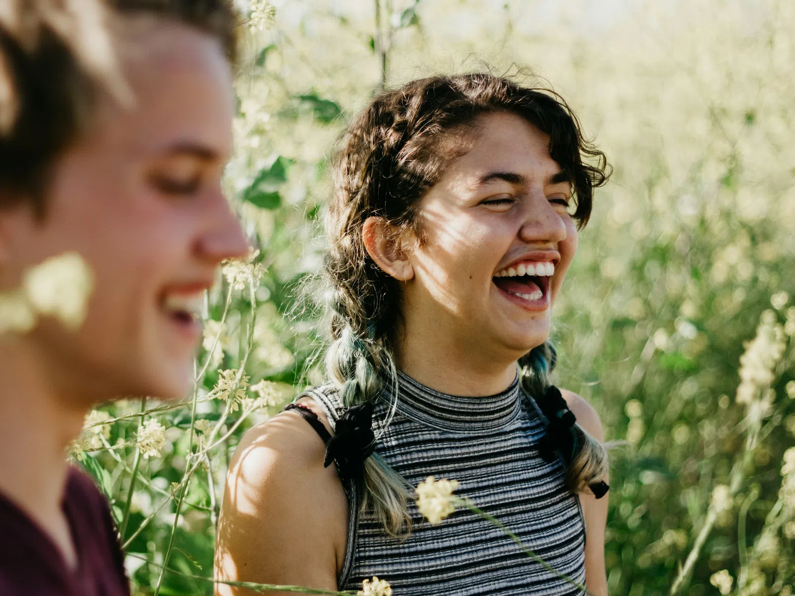 Two young people laughing outdoors in a sunlit natural field with green plants in the background