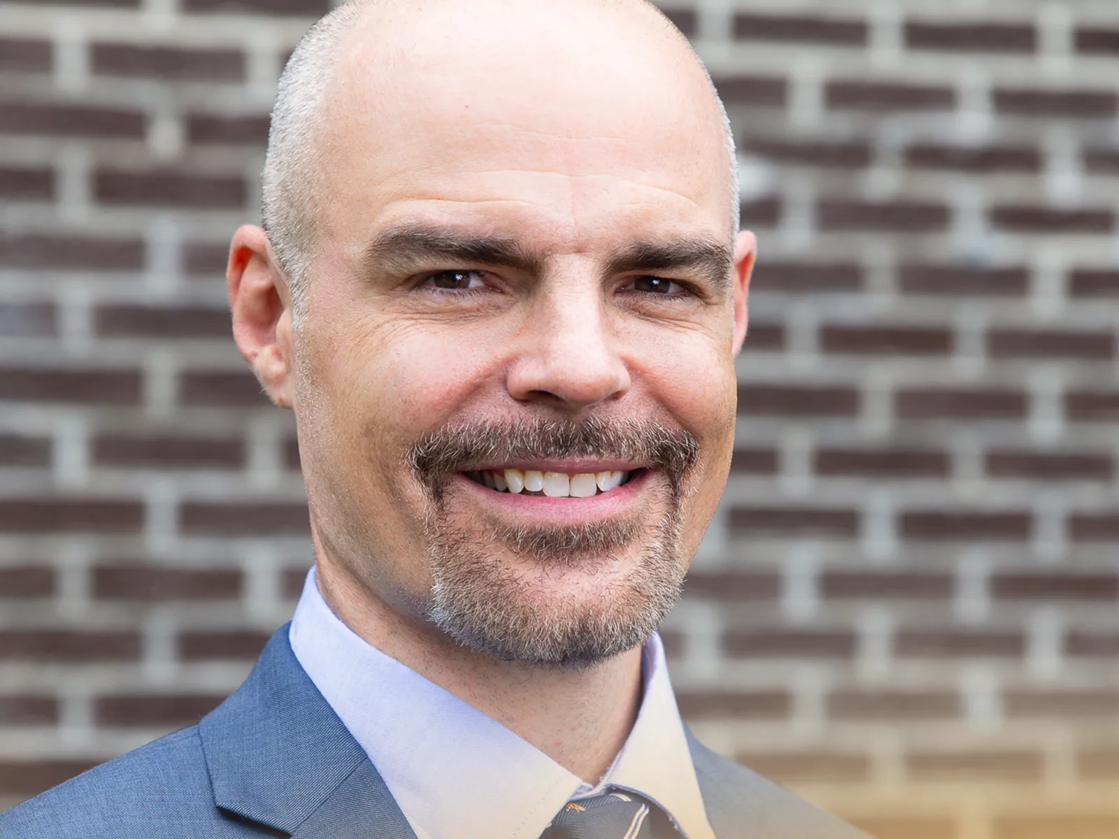 Smiling bald man with a goatee wearing a blue suit and tie standing in front of a brick wall outdoors