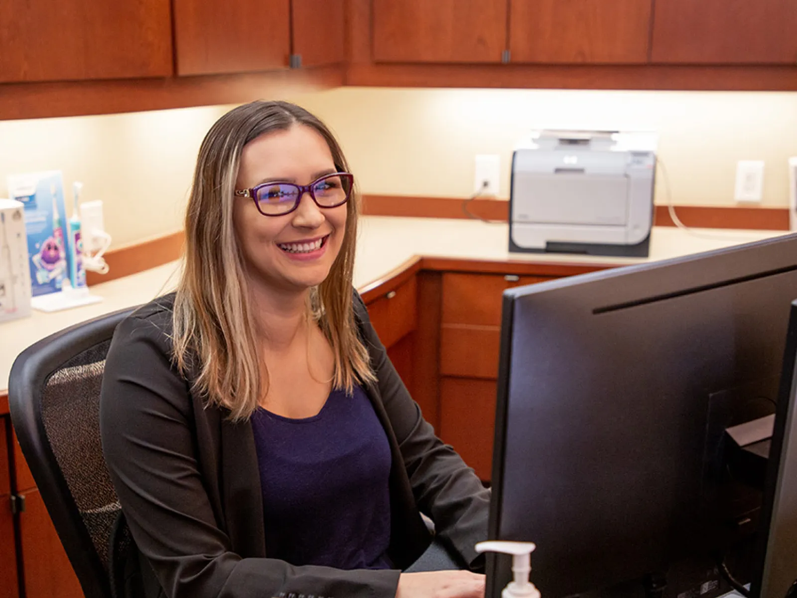 Smiling woman with glasses working at a computer in a modern office with wood cabinets and hand sanitizer bottles.