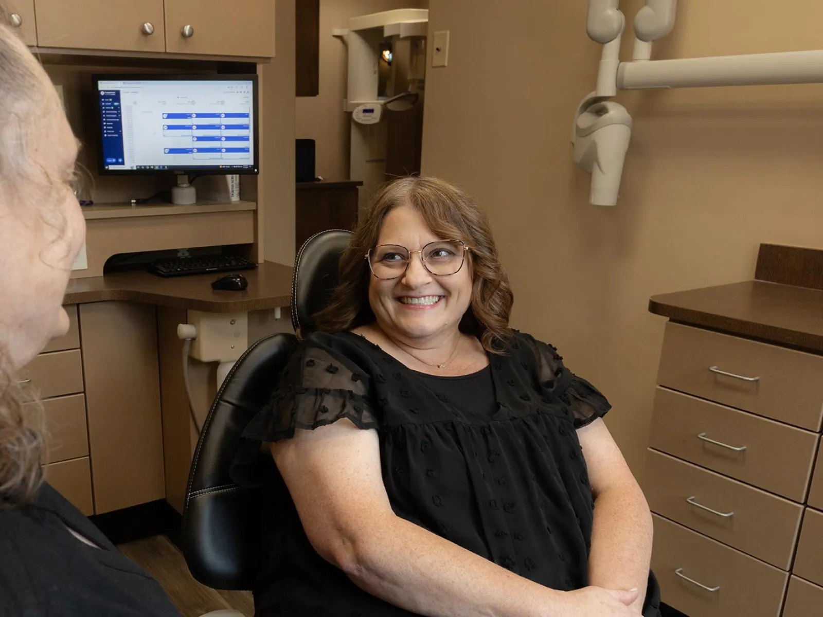 Smiling woman sitting in dental chair talking with another woman in dental office with computer screen.
