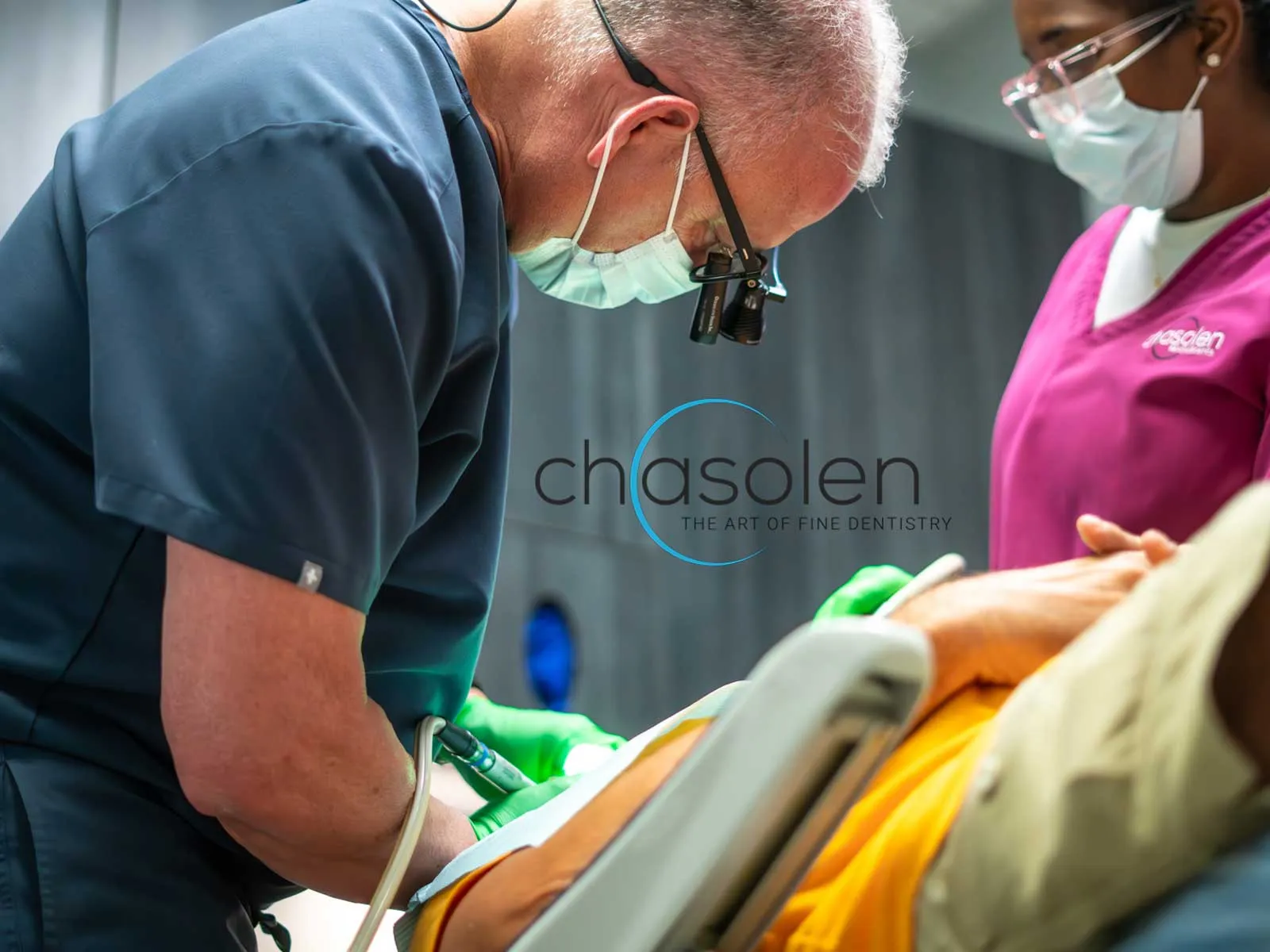 Dentist wearing magnifying glasses and mask treating patient with assistant in dental clinic