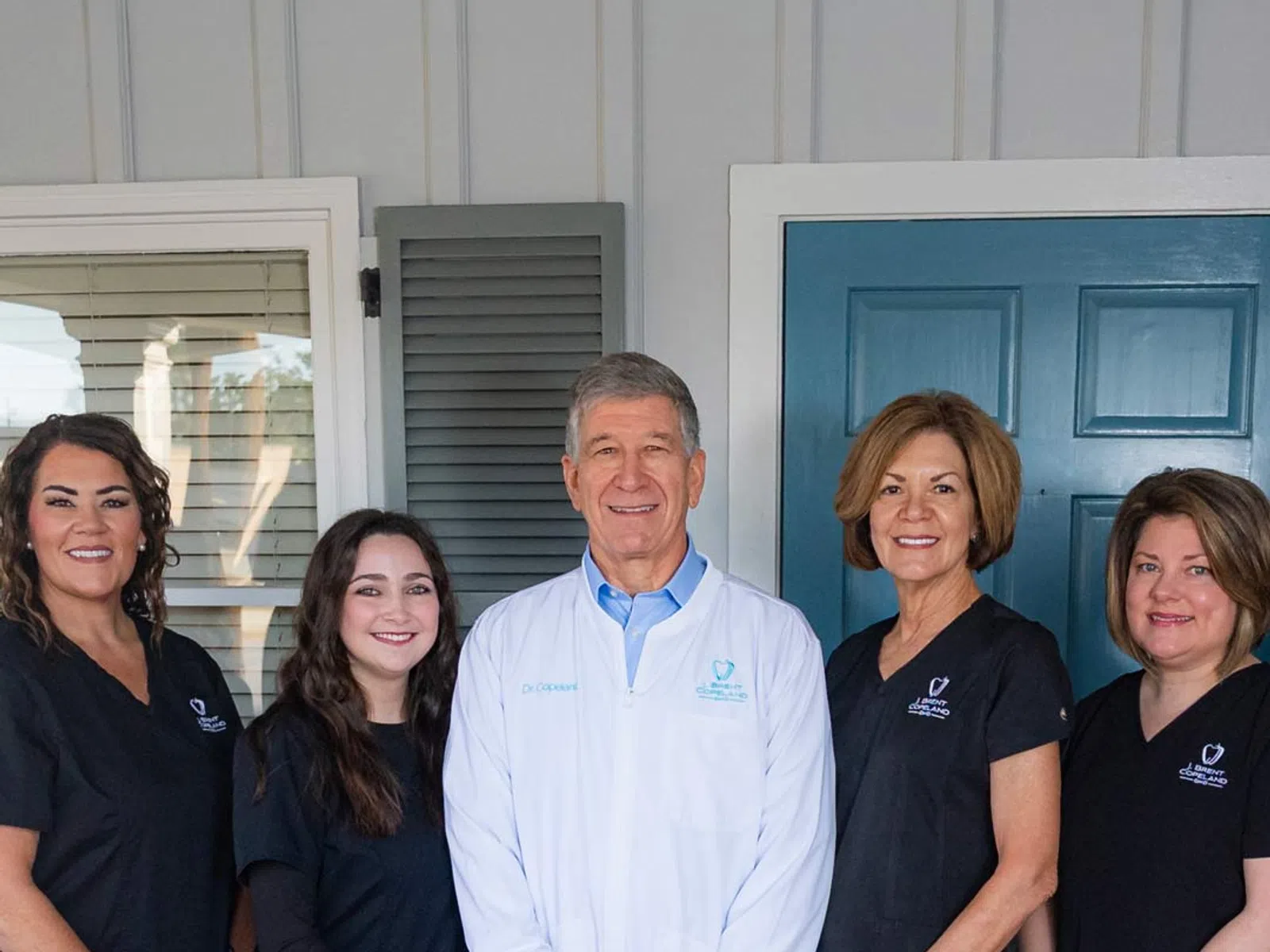 Dental team of five professionals standing on a tiled porch in front of blue door smiling at camera.