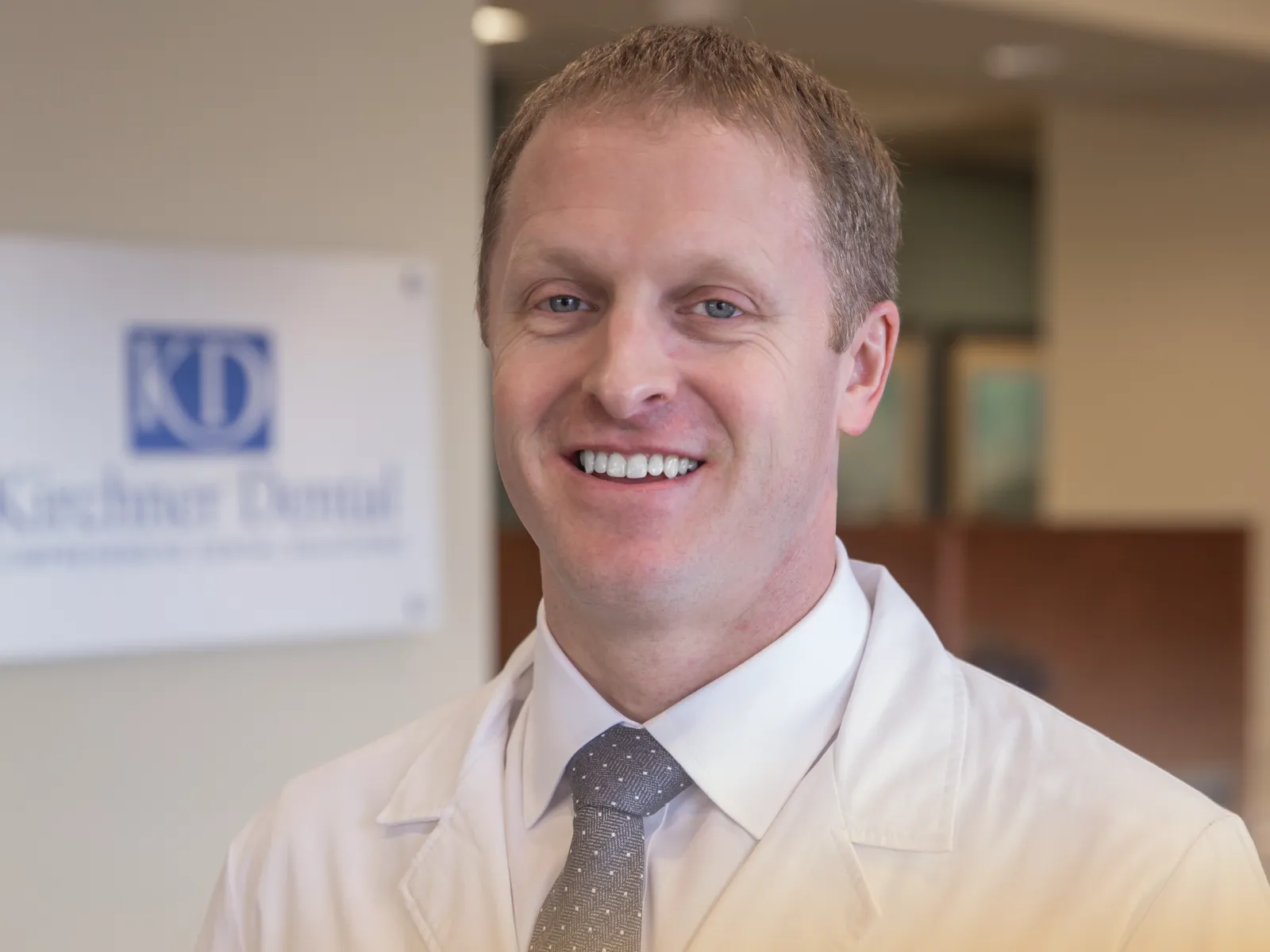 Smiling male dentist in white coat standing in dental office with Kiechner Dental sign in background