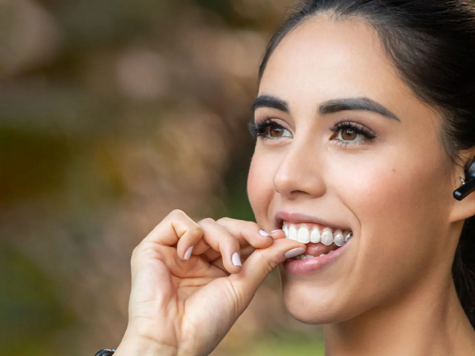 Young woman smiling as she fits clear dental aligners outdoors wearing a fitness watch and earbud.