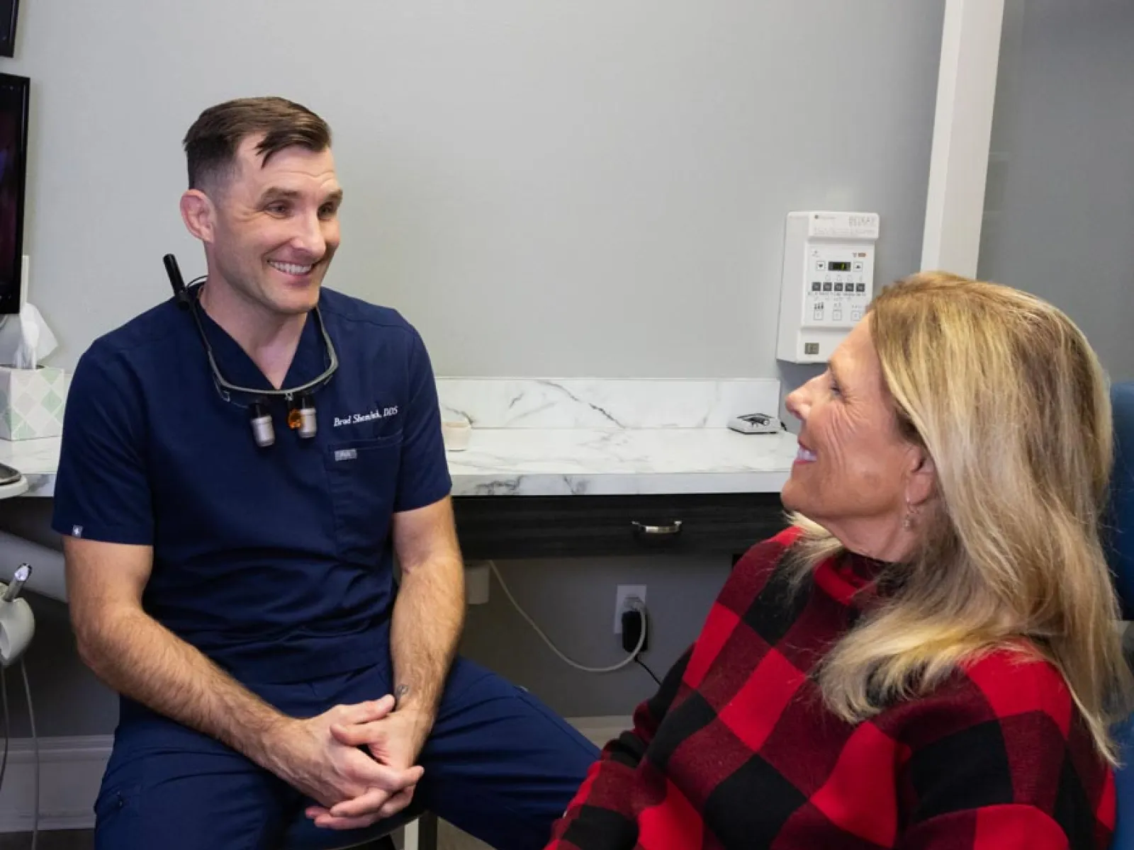 Dentist in navy scrubs smiling and talking with a female patient in a dental office consultation.
