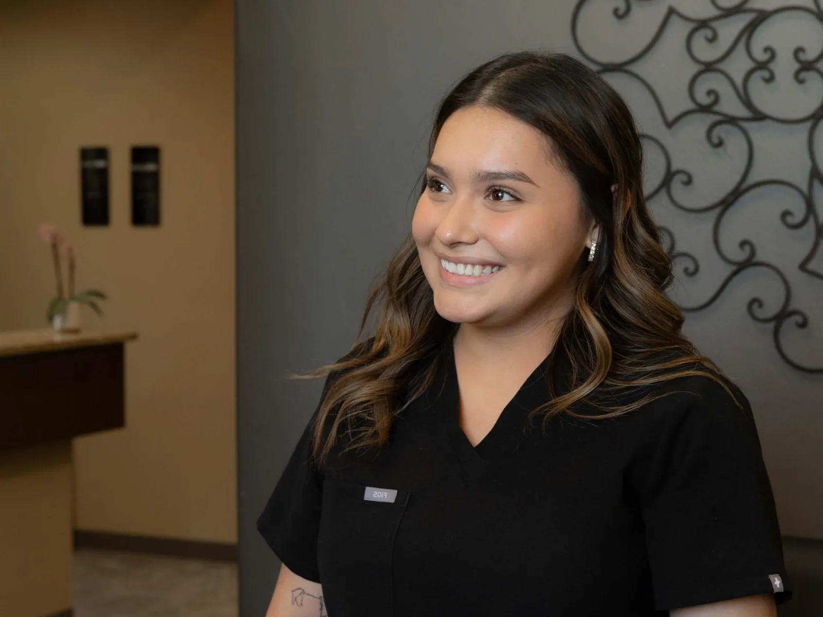 Smiling woman in black scrubs standing against a gray wall with decorative ironwork, looking right.