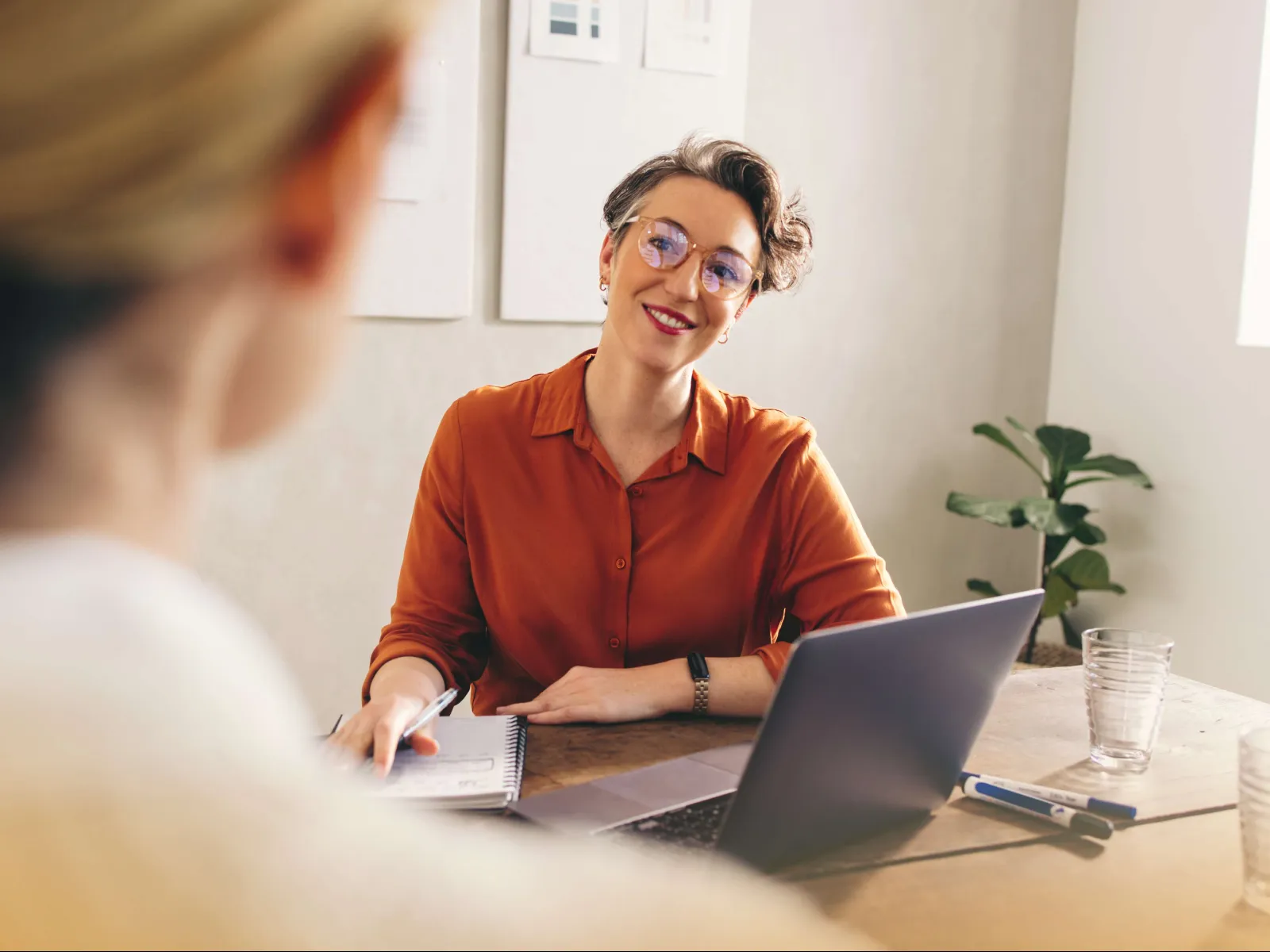 Woman in orange shirt smiling and talking while working on laptop during a meeting in bright office.