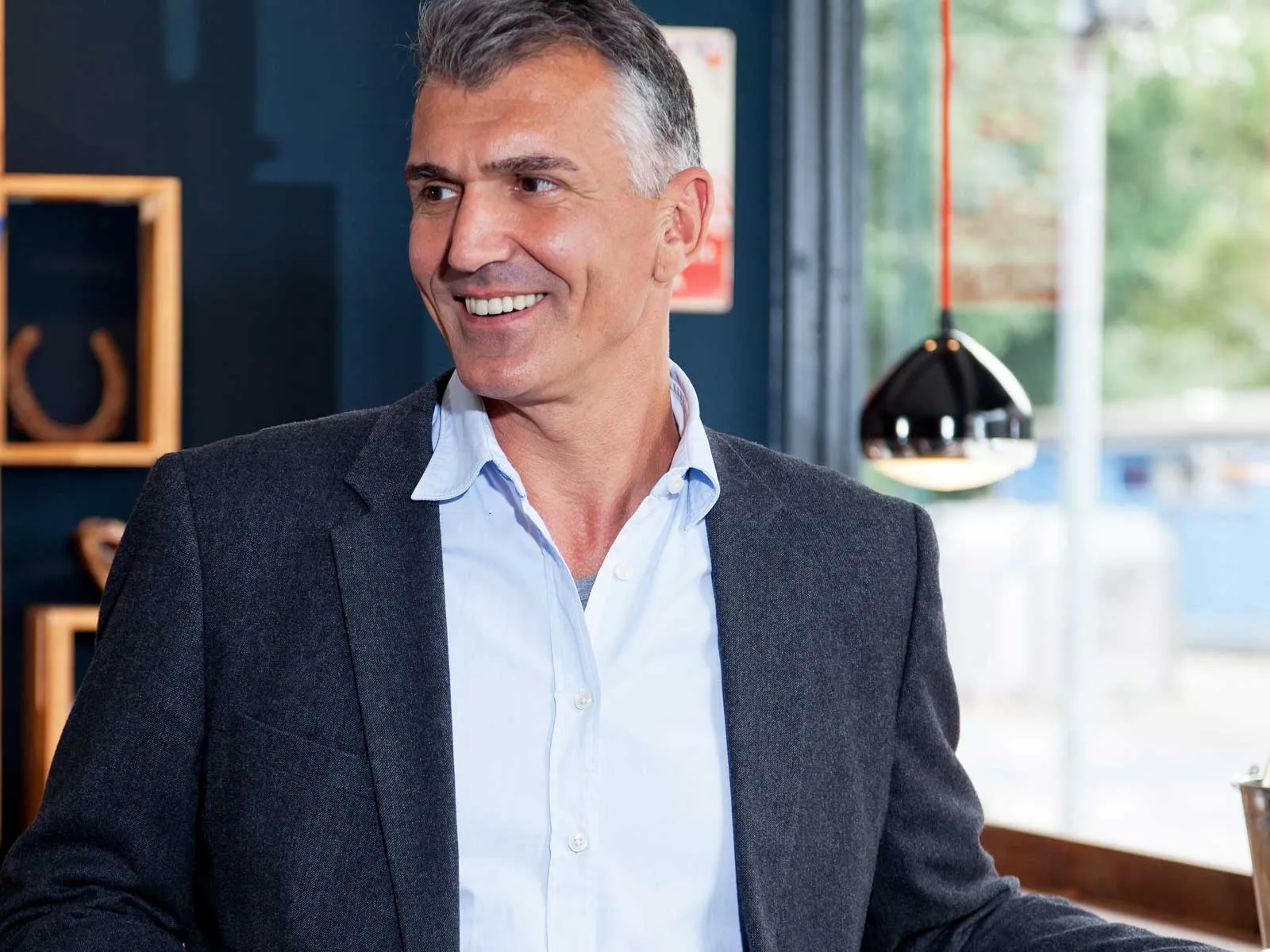 Smiling mature man in a gray blazer and blue shirt standing indoors near a window with modern decor.