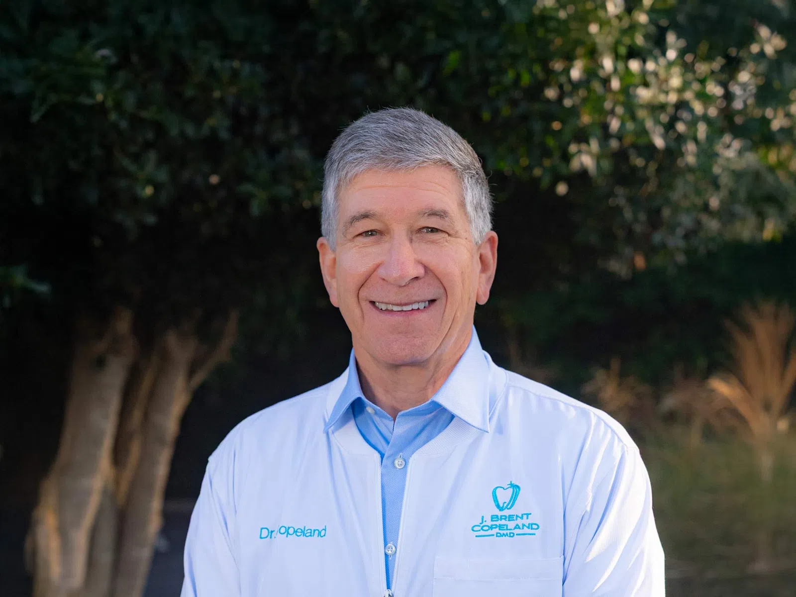 Smiling male dentist wearing white coat with dental logo standing outdoors with trees in background