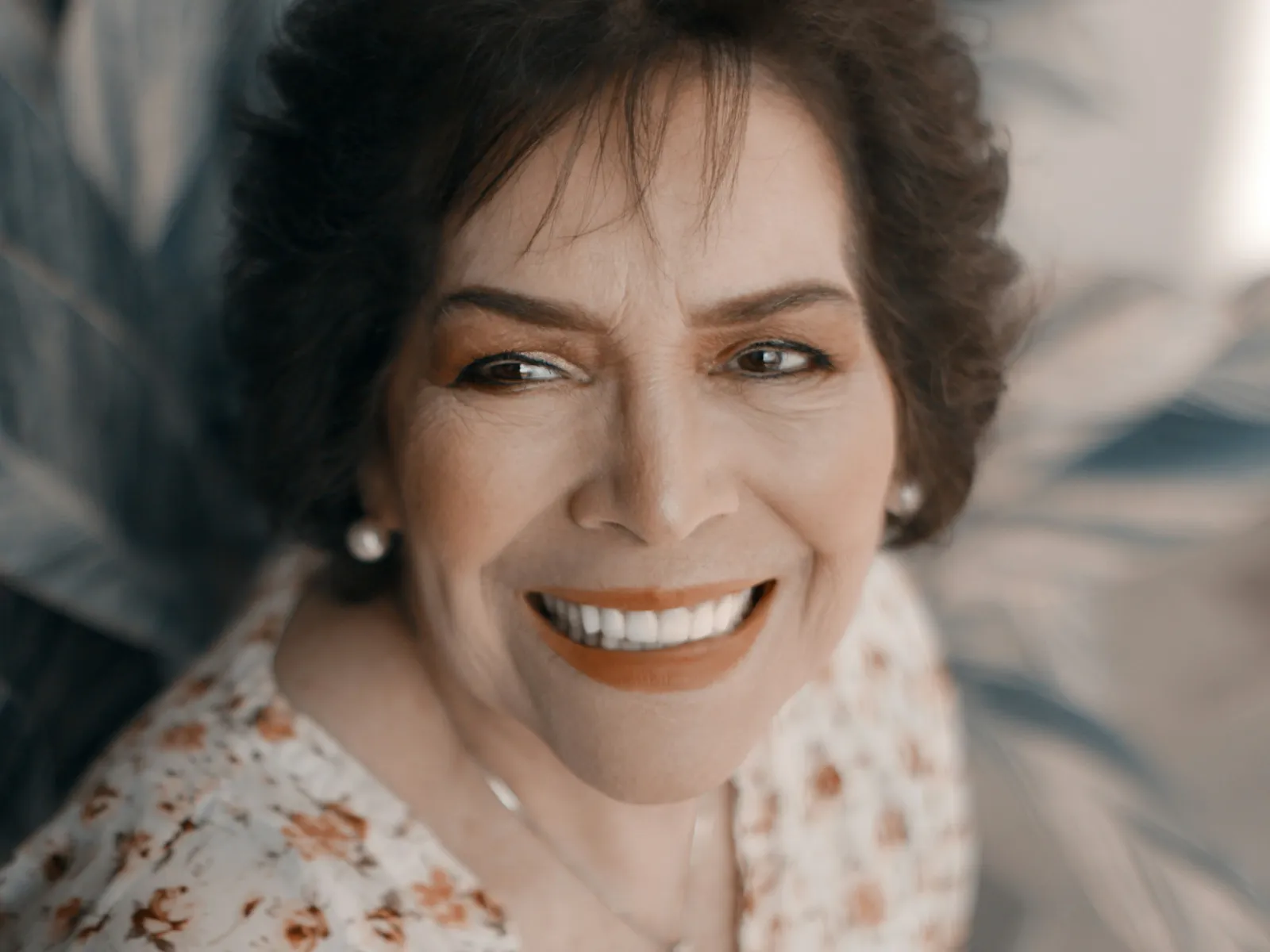 Smiling mature woman with short dark hair wearing floral blouse and pearl earrings in soft natural light.