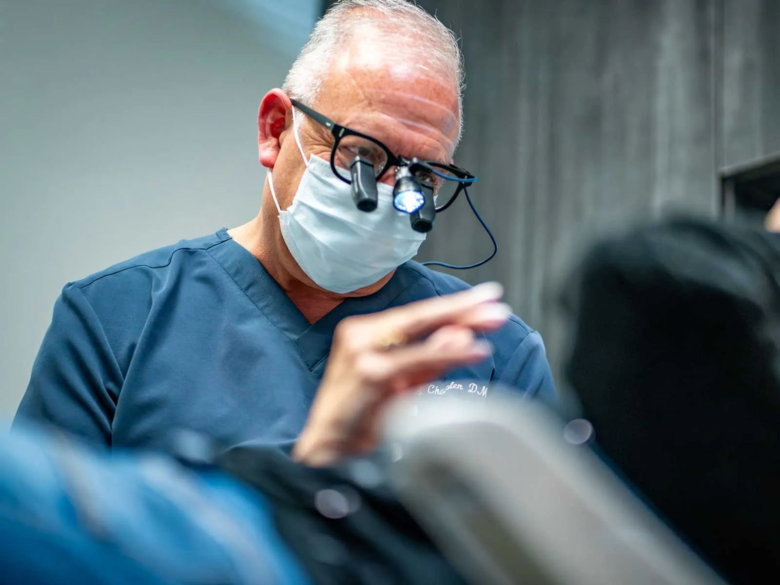 Dentist wearing magnifying glasses and mask examines patient in a clinical setting with focused lighting.