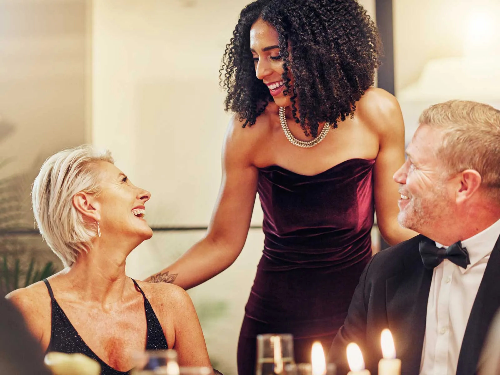 Smiling woman in elegant velvet dress chatting with two formally dressed guests at a cozy dinner party.