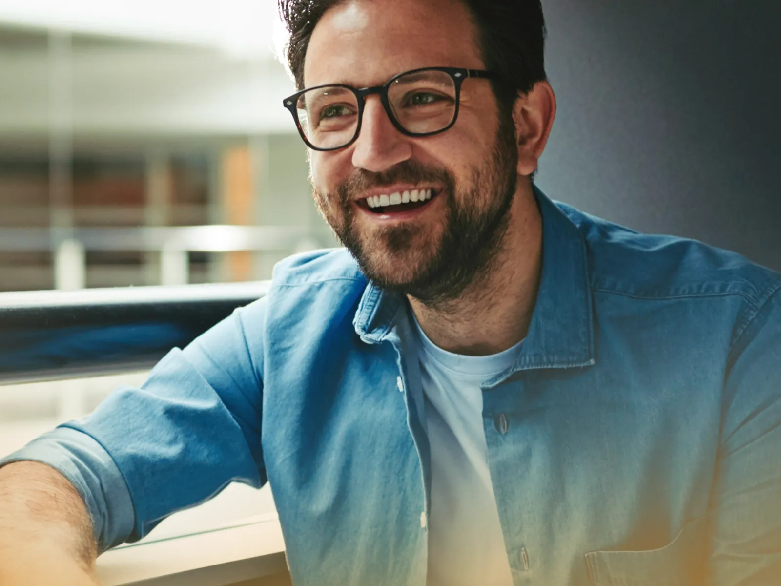 Smiling man wearing glasses and a blue denim shirt, sitting indoors in a bright modern space.