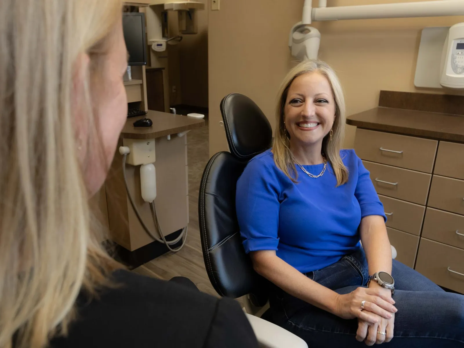 Smiling woman in blue shirt sitting in dental chair talking to another person in dental office.