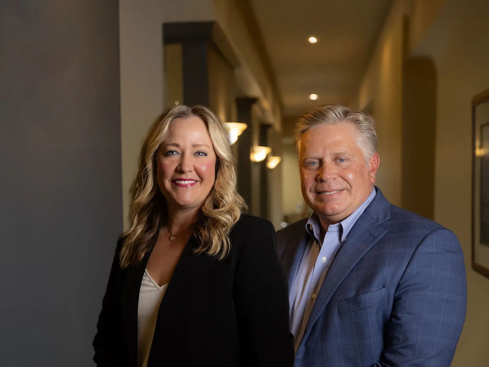 Professional middle-aged man and woman smiling in an office hallway with warm lighting.