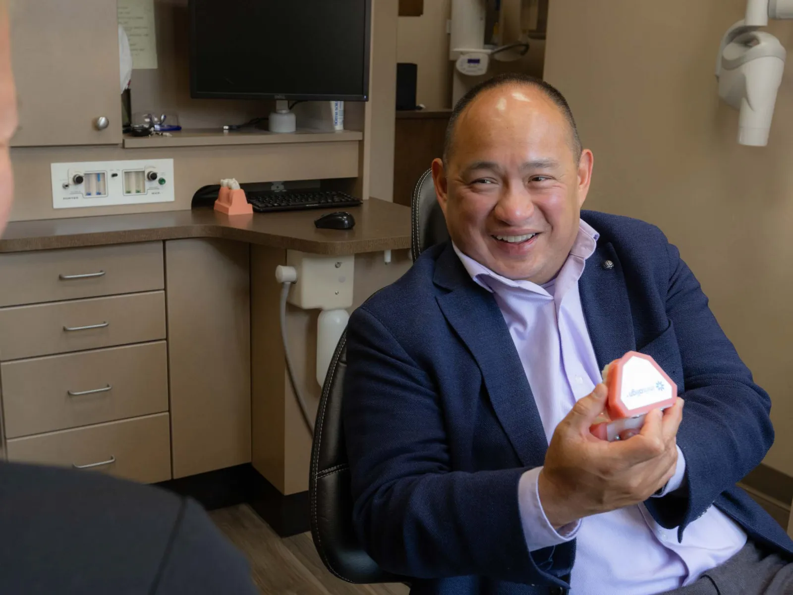 Dentist in office showing dental mold to patient during consultation with chair and equipment in background