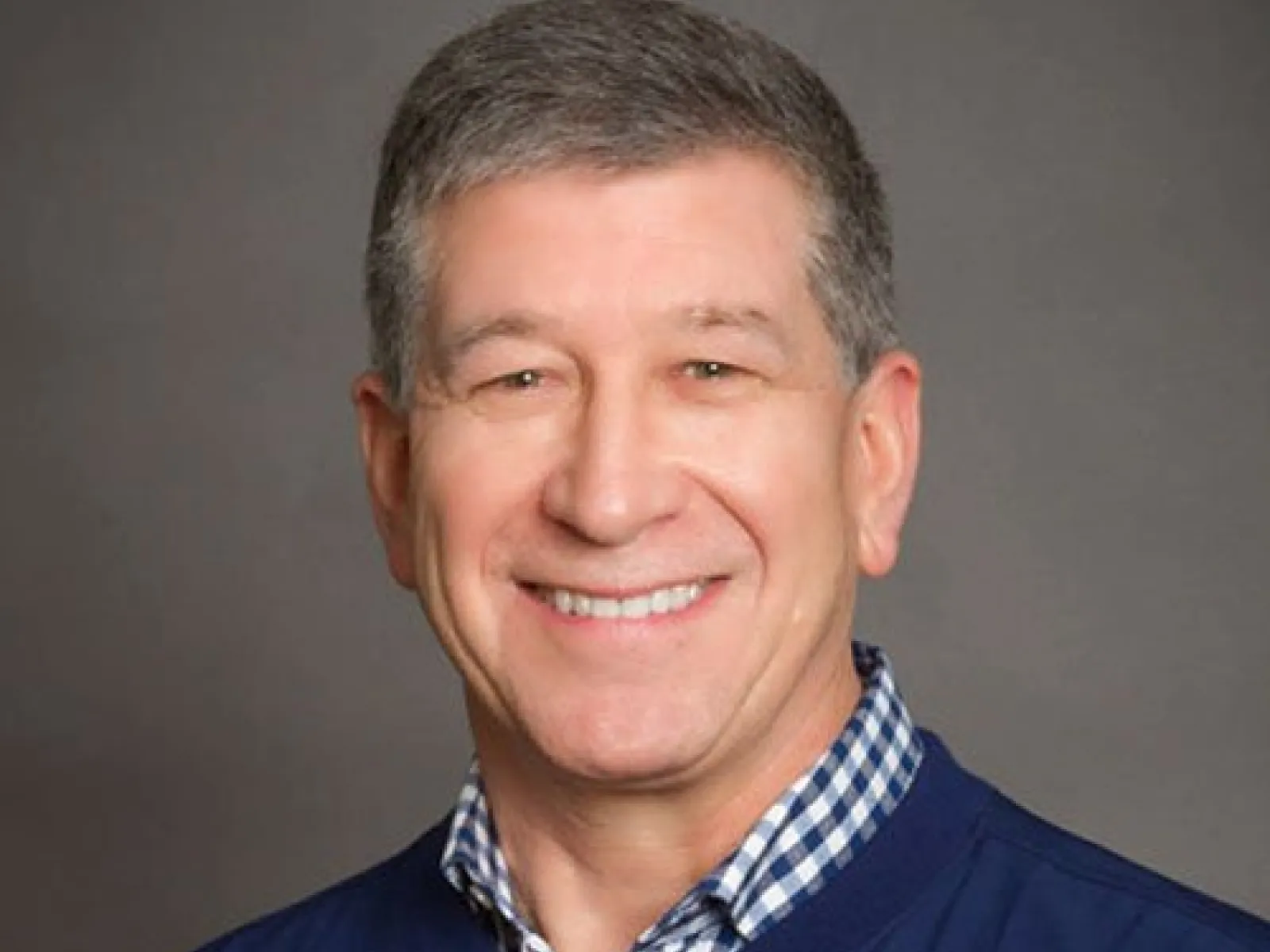 Smiling middle-aged man wearing a navy blue jacket and checkered shirt against a gray background