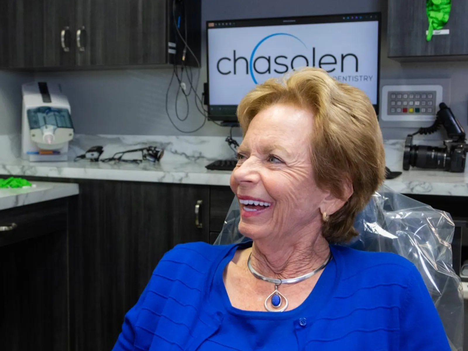 Smiling elderly woman wearing blue seated in a dental office with Chasalen Dentistry logo on screen behind her