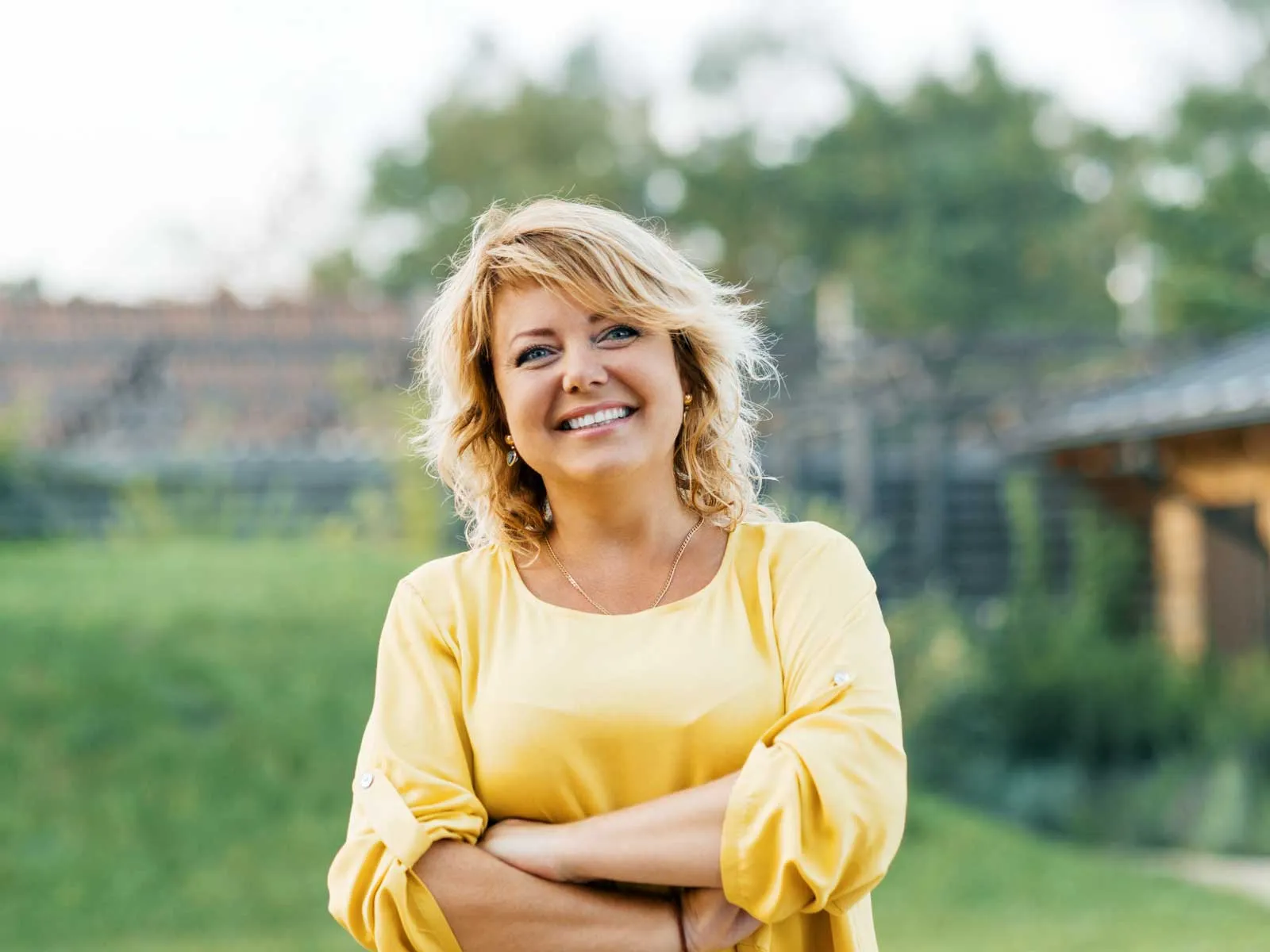 Smiling woman with blonde hair wearing a yellow blouse standing outdoors with arms crossed and blurred background