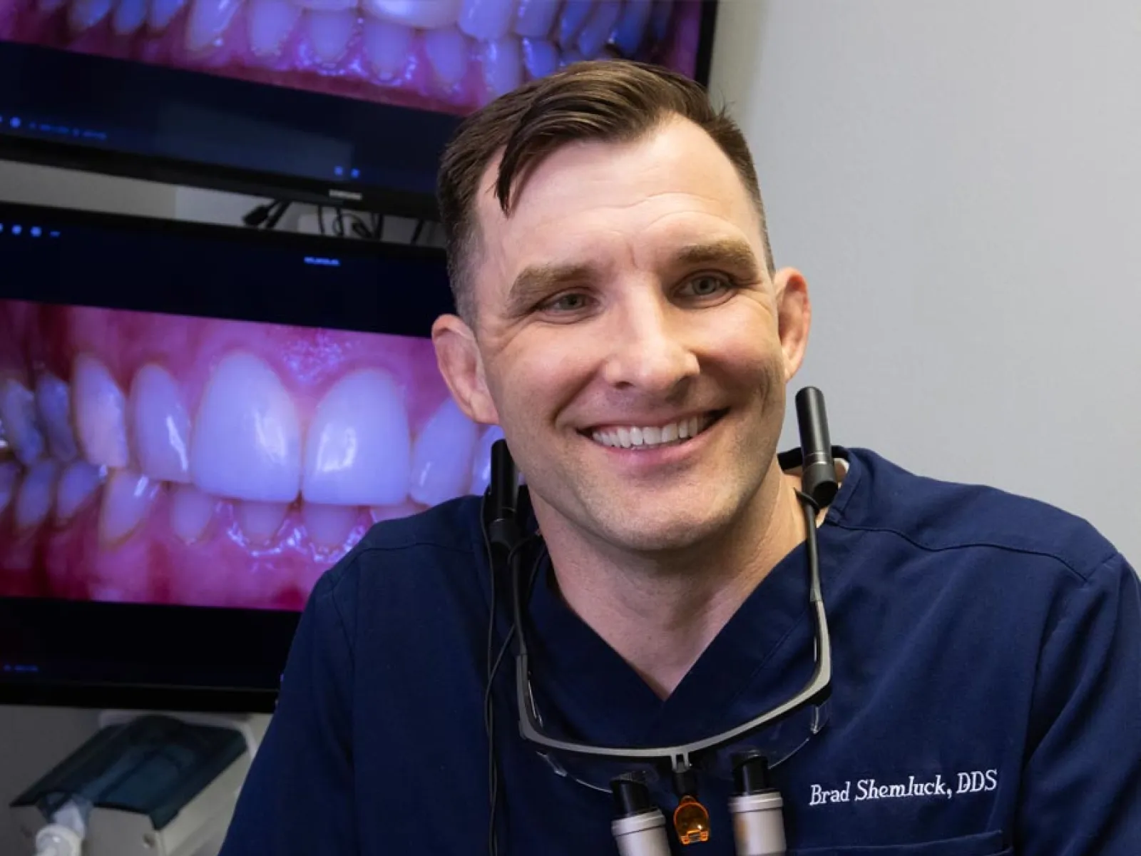 Smiling male dentist in navy blue scrubs with dental tools, screens showing close-up of teeth behind him