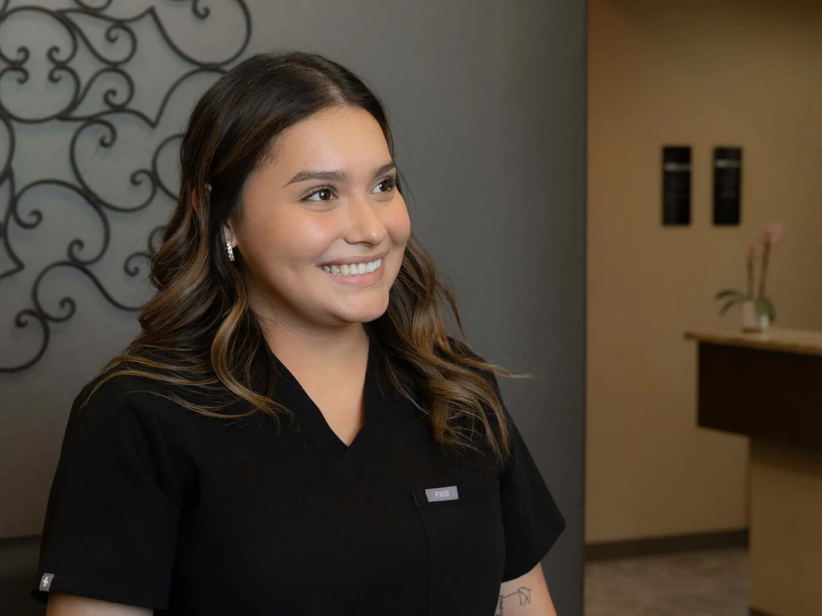 Smiling woman in black scrubs with long hair standing indoors near decorative wall and counter with flower vase.