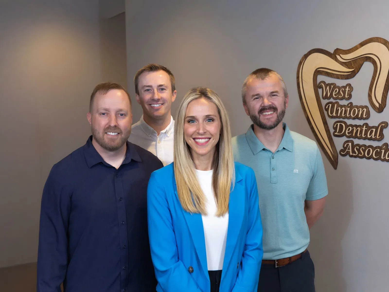 Four dental professionals smiling inside West Union Dental Associates office with logo on wall.