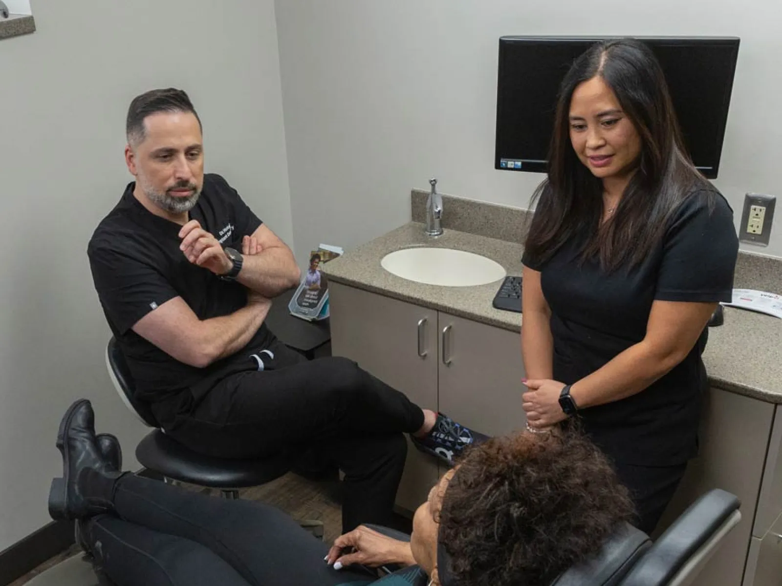 Two healthcare professionals consult with a patient reclining in a medical office chair.