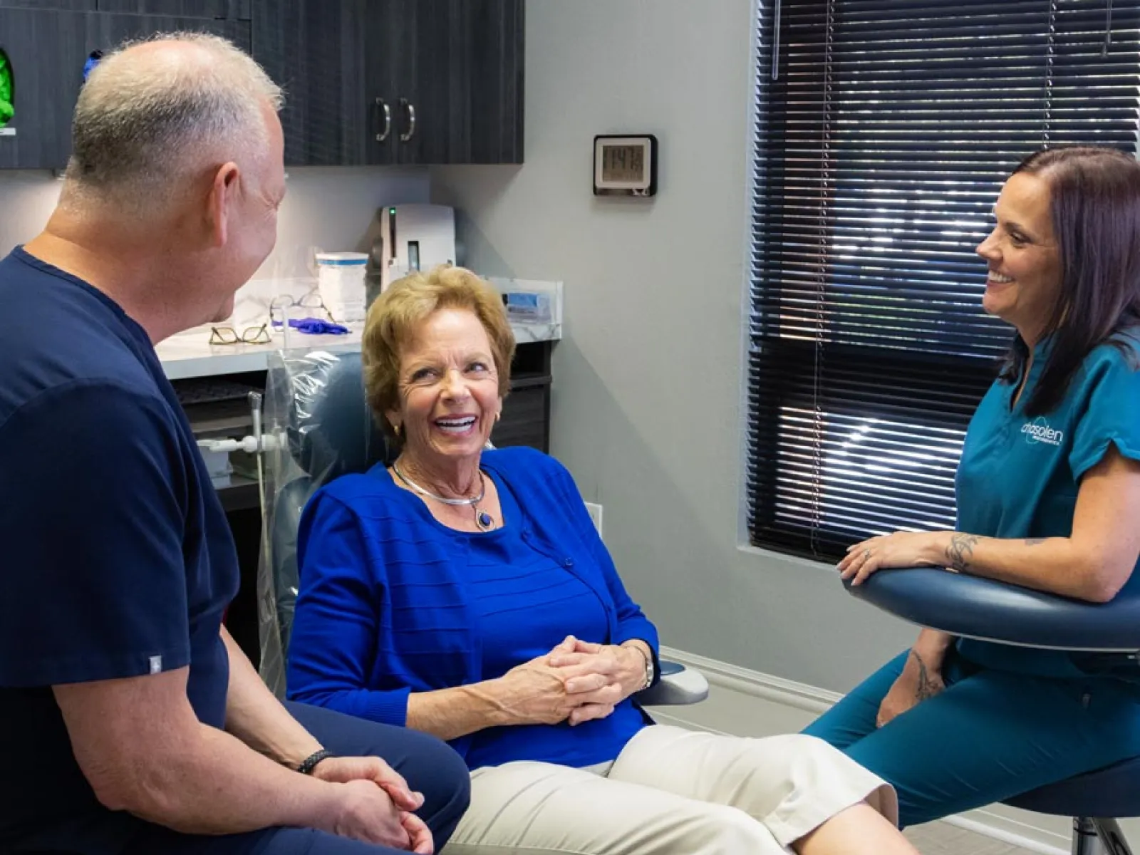 Senior woman smiling in dental chair talking with two dental professionals in modern clinic room