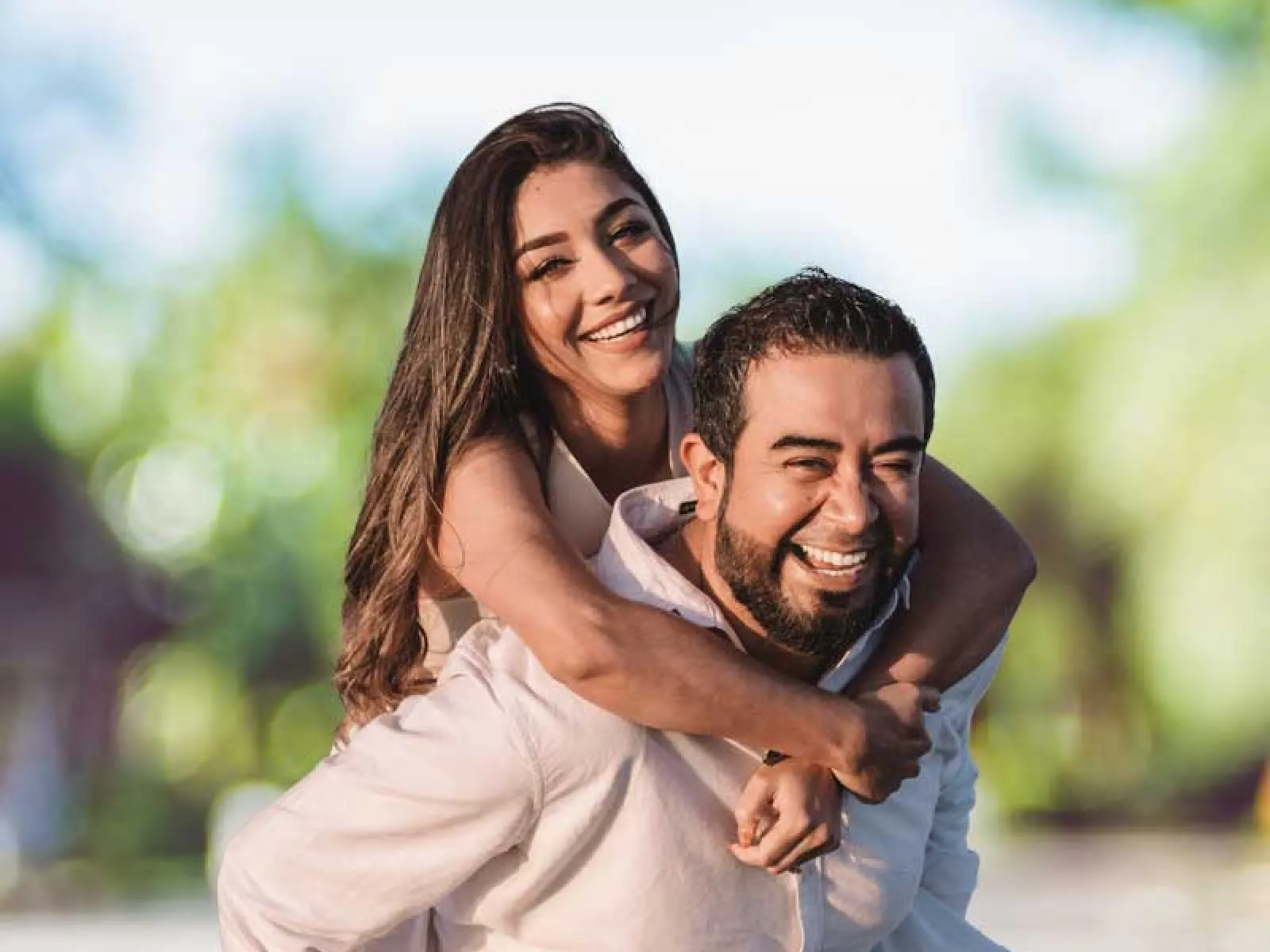 Happy couple outdoors, woman giving piggyback ride to smiling man with blurred green background