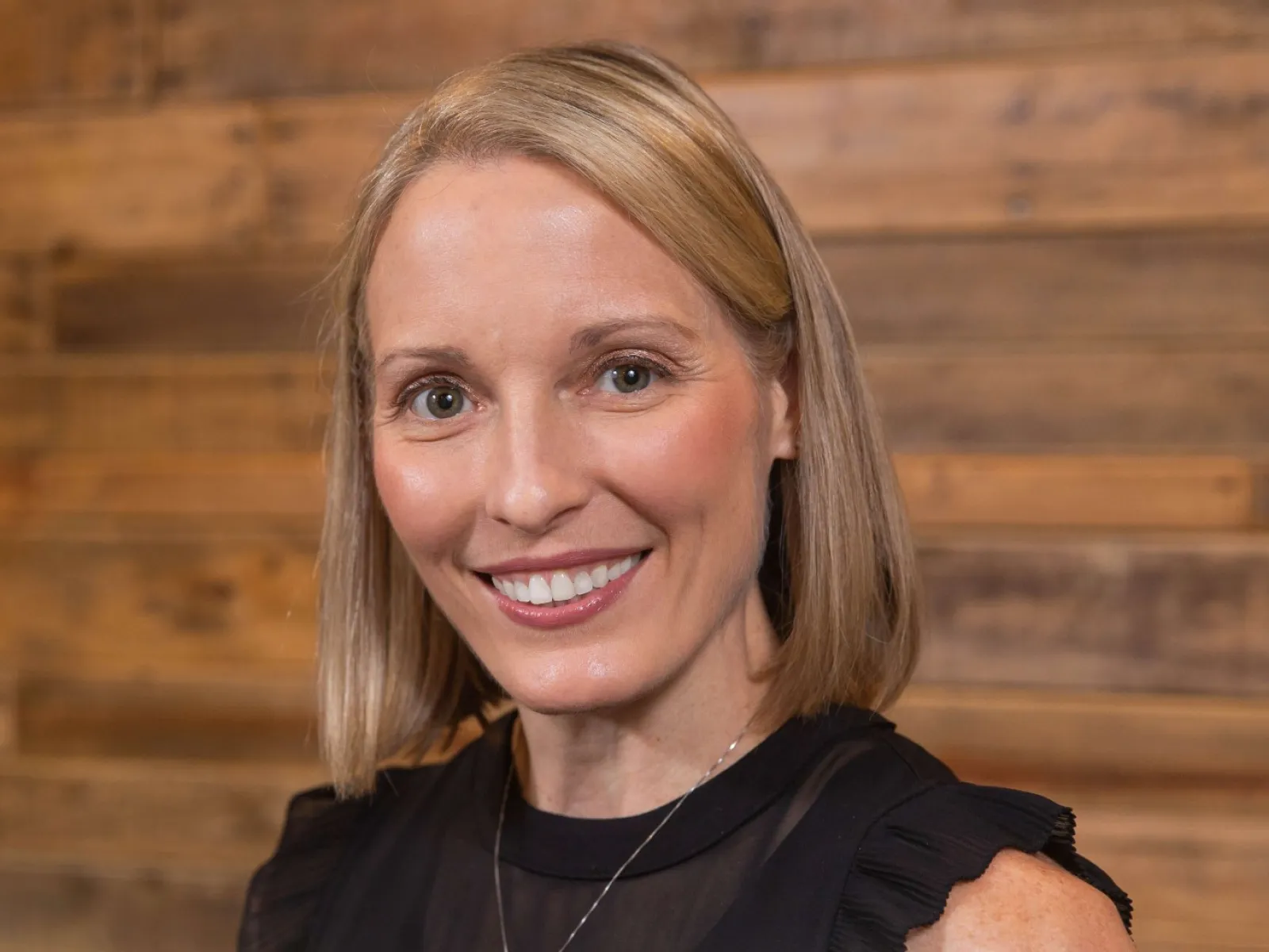 Smiling blonde woman with short hair wearing a black ruffled sleeveless top in front of wooden wall.