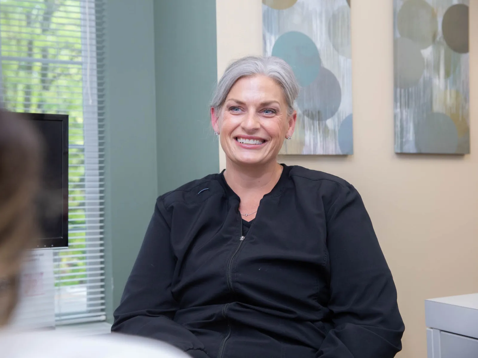 Smiling middle-aged woman with gray hair wearing black jacket sitting indoors in a bright room.