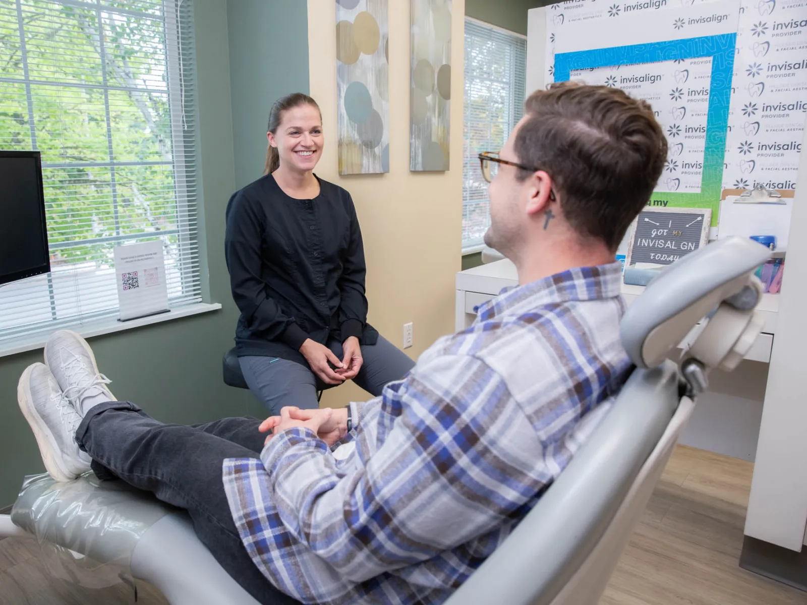 Patient sitting in dental chair talking with smiling dental professional in a bright office with window and Invisalign signs.