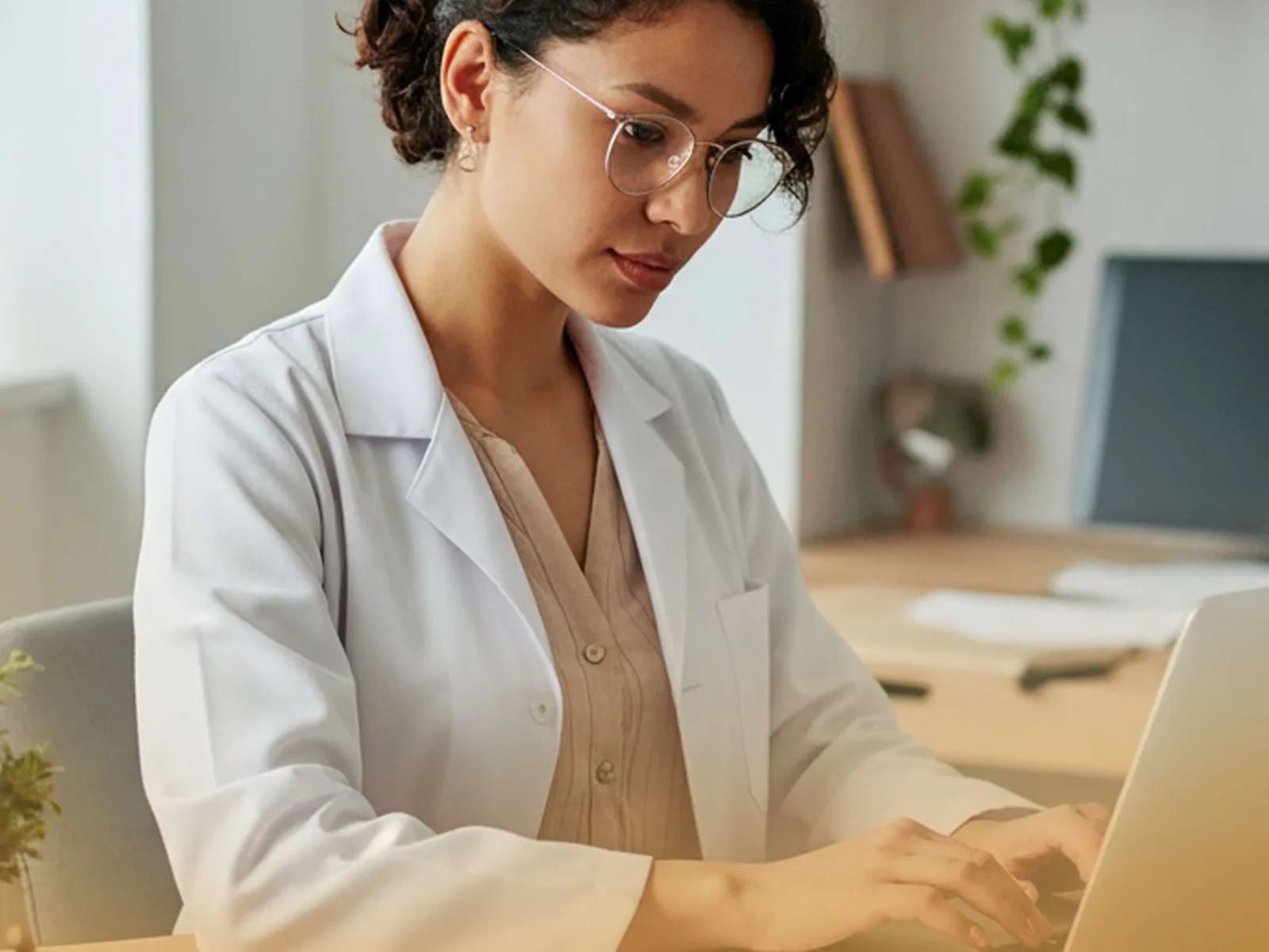 Female doctor wearing glasses and a lab coat typing on a laptop in a bright, organized office space.