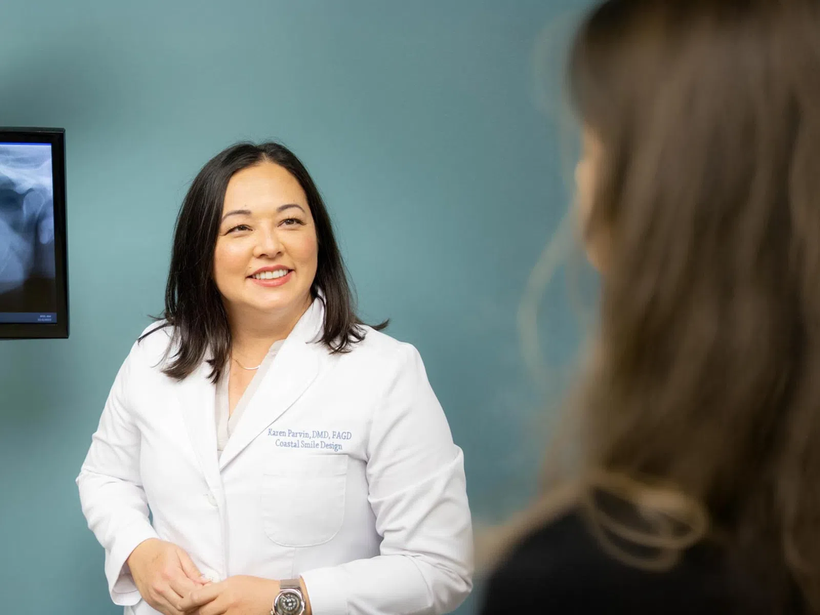 Female dentist in white coat smiling while talking to a patient with dental X-ray visible on screen