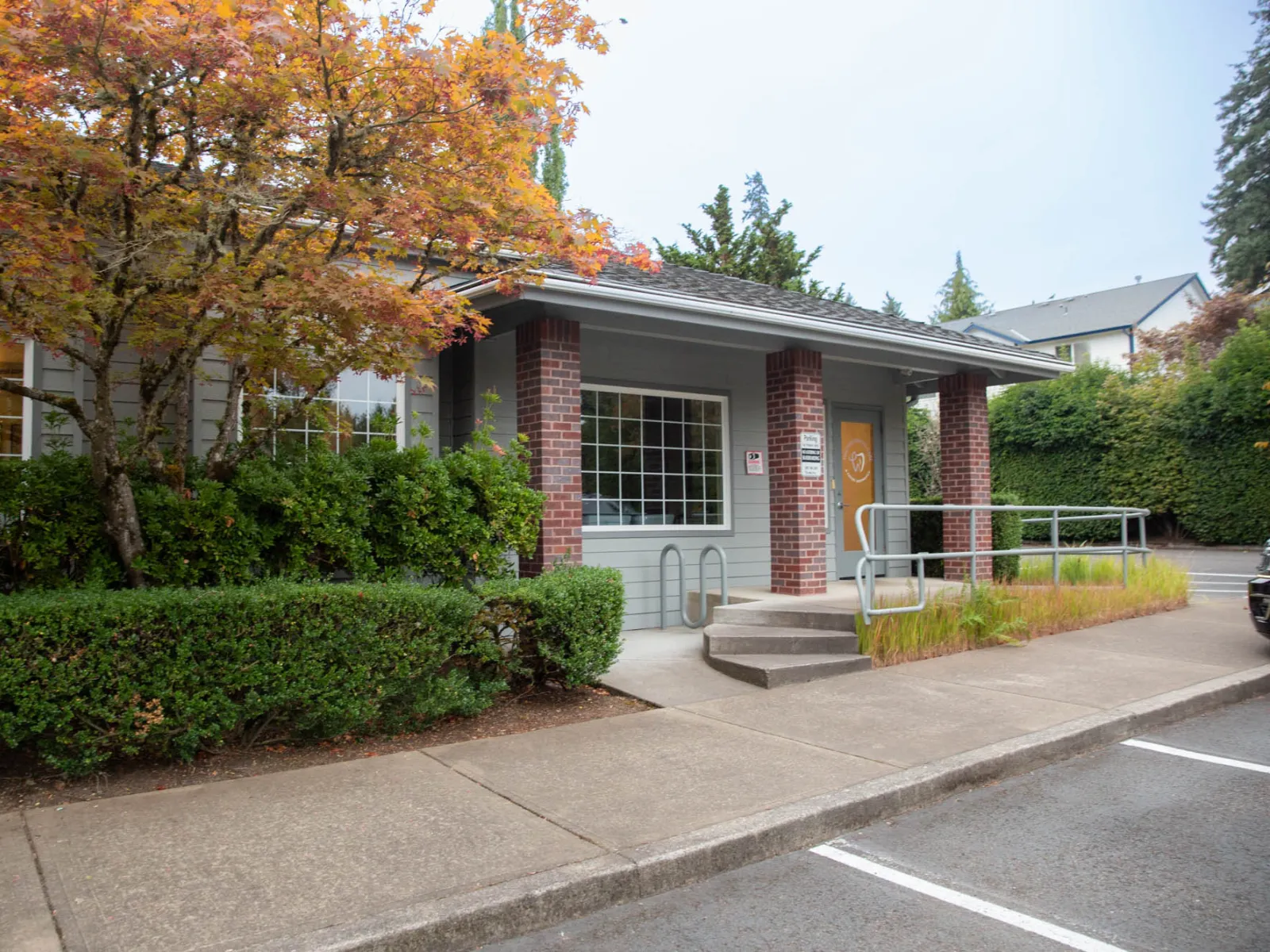 Small building entrance with brick pillars, wheelchair ramp, and autumn foliage beside sidewalk and parking lot.