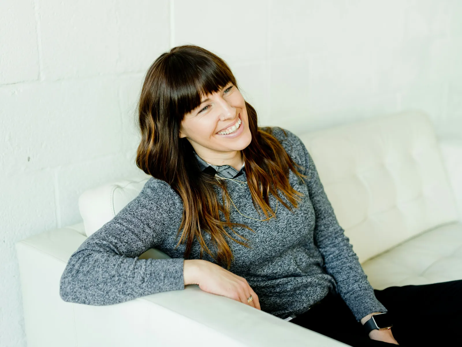 Smiling woman with brown hair wearing a gray sweater sitting relaxed on a white couch against a white wall.