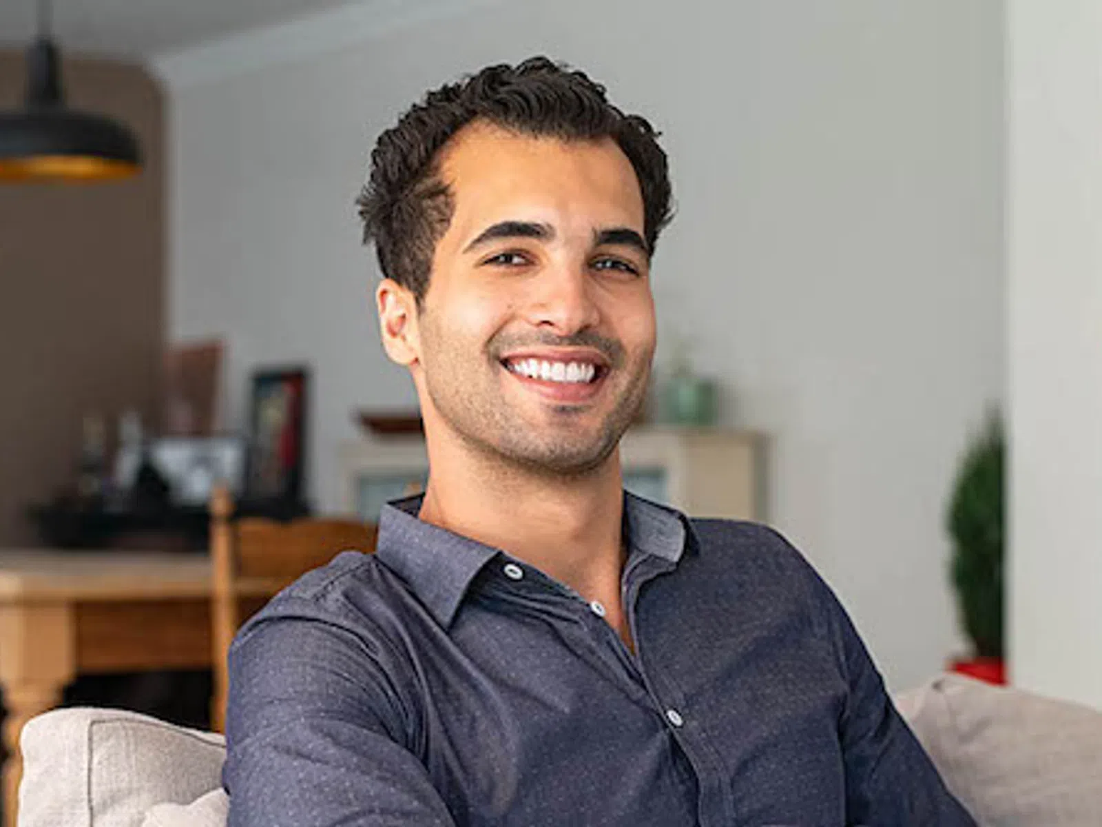 Smiling young man with dark hair and gray shirt sitting on a couch in a modern living room.