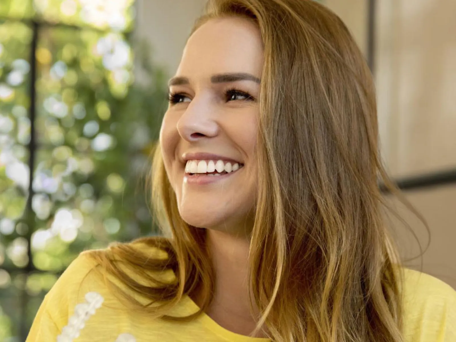 Smiling woman in yellow shirt holding a clear dental aligner with long light brown hair indoors.