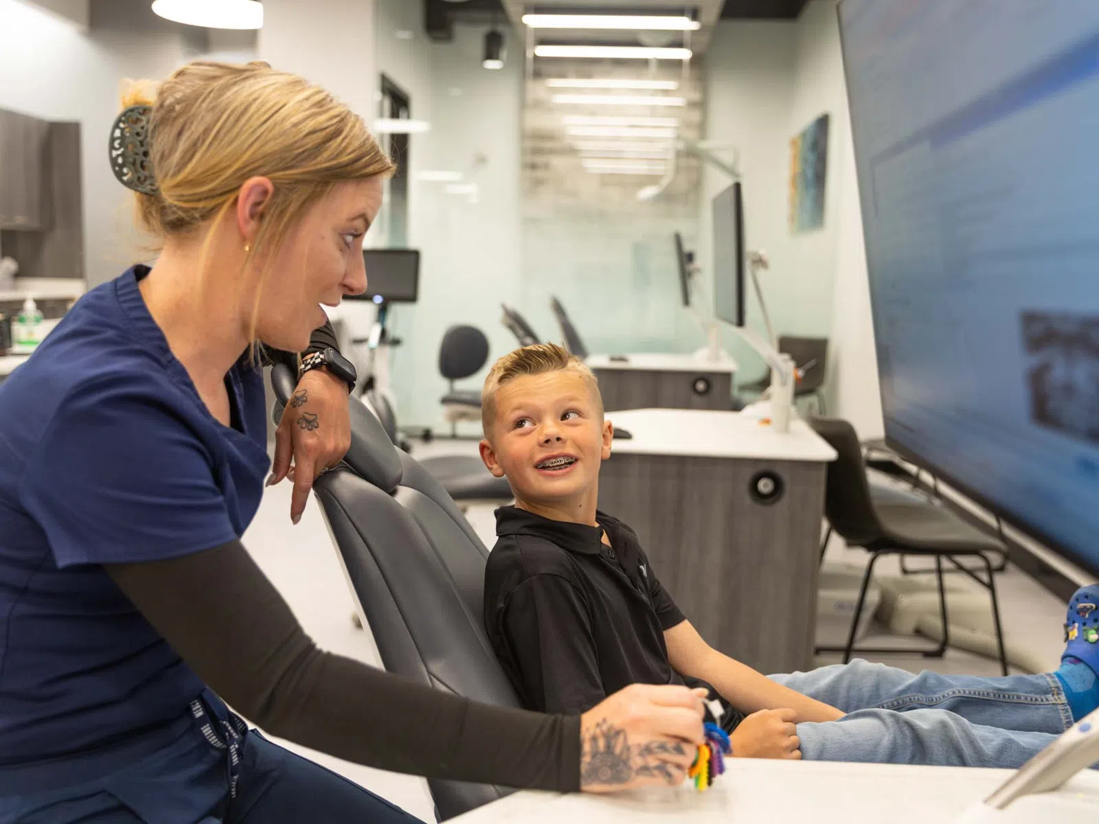 Child sitting in dental chair smiling at female dentist in modern clinic with computer screen.