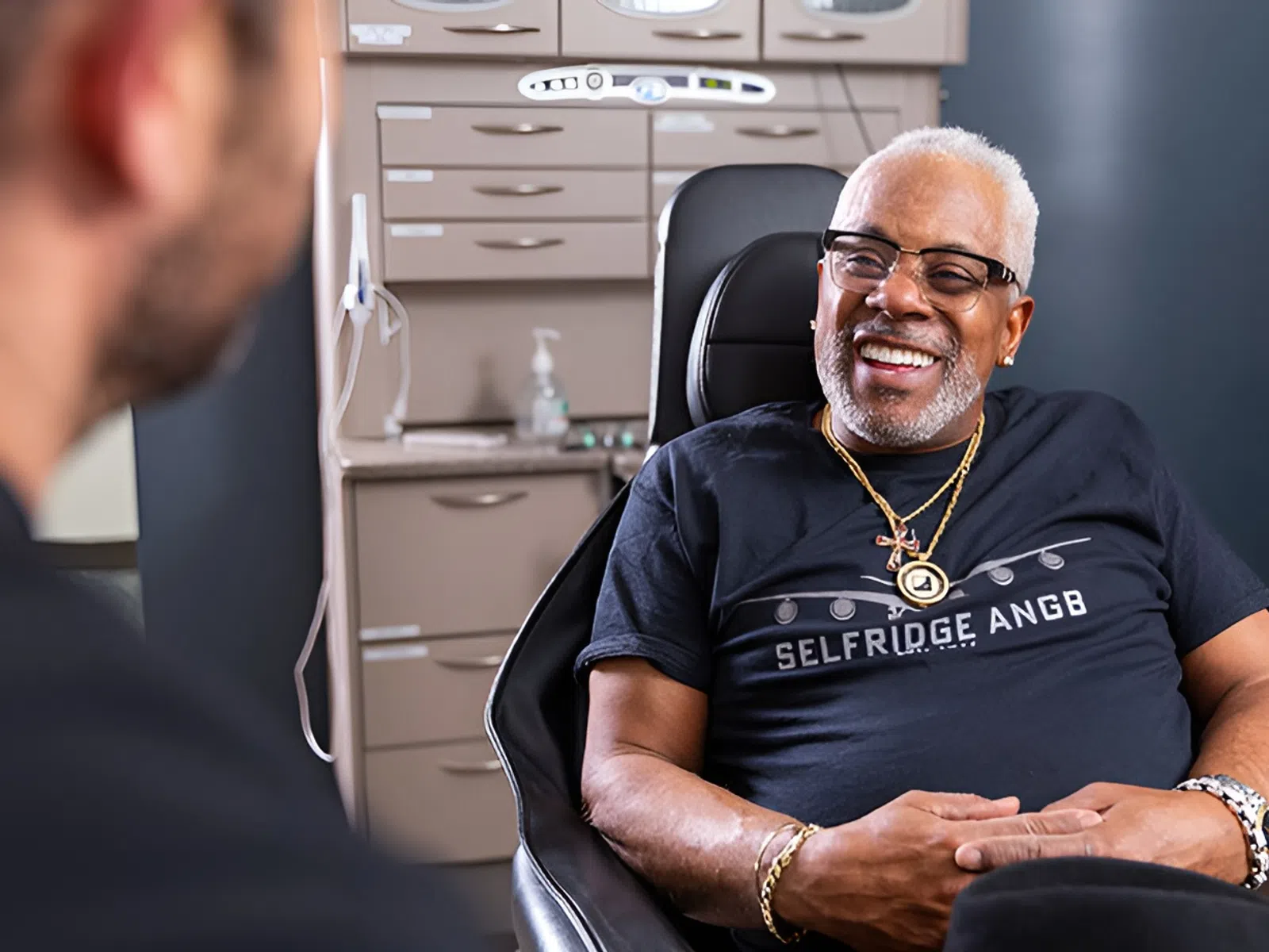 Smiling elderly man wearing glasses and jewelry sitting in a chair during a consultation in a modern office.