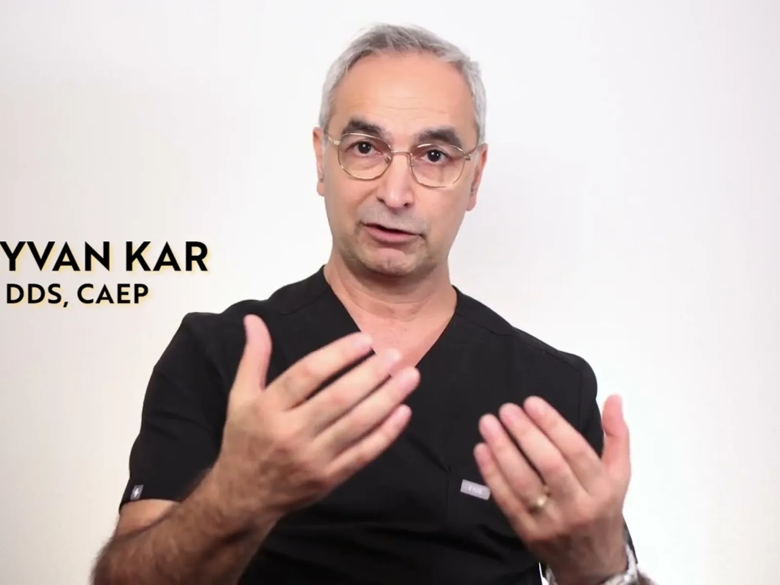Male dentist Keyvan Kar in black scrubs explaining with hand gestures against white background.