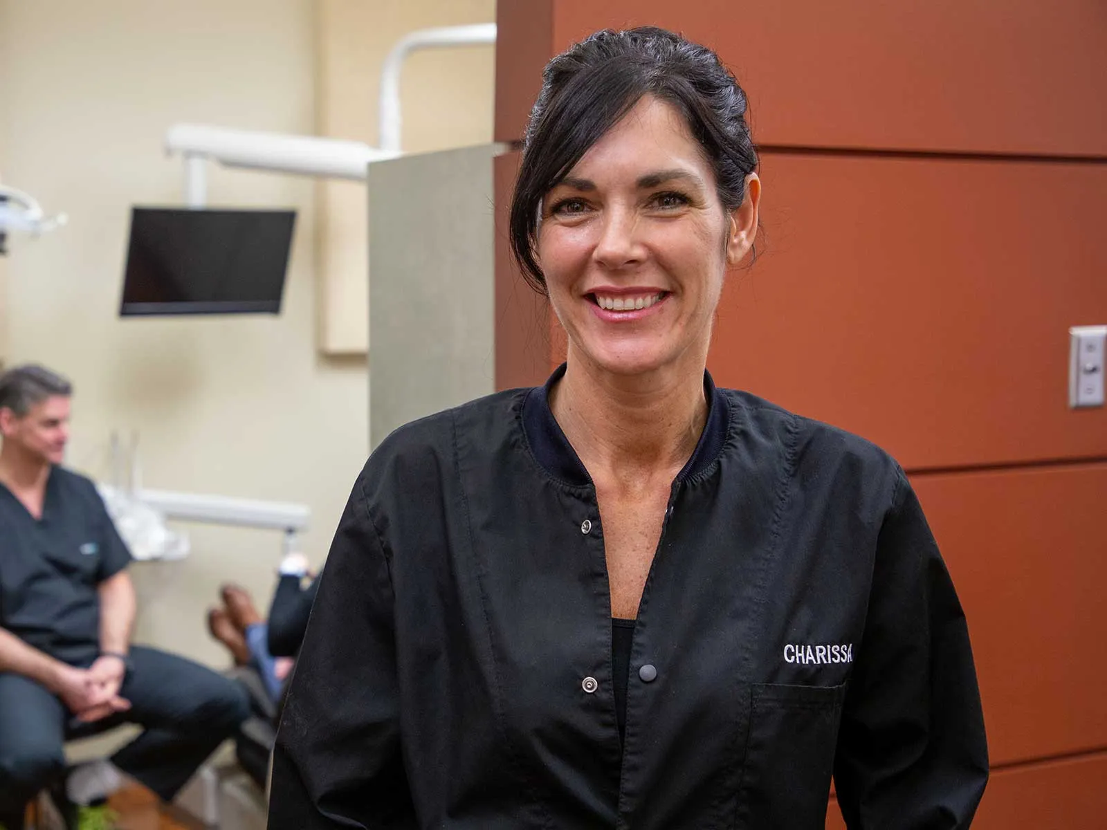 Smiling female dental professional in black scrubs in a dental office with a colleague in the background