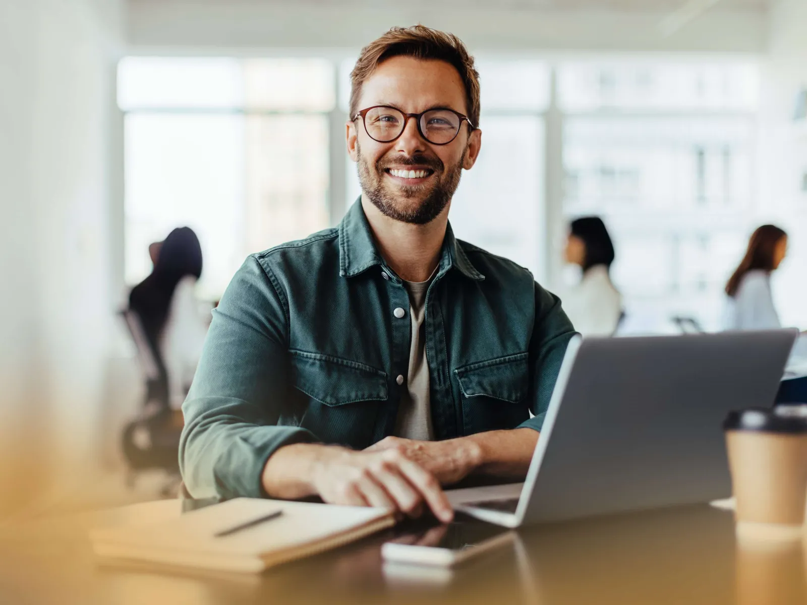 Smiling man with glasses working on a laptop at a modern office desk with blurred coworkers in background