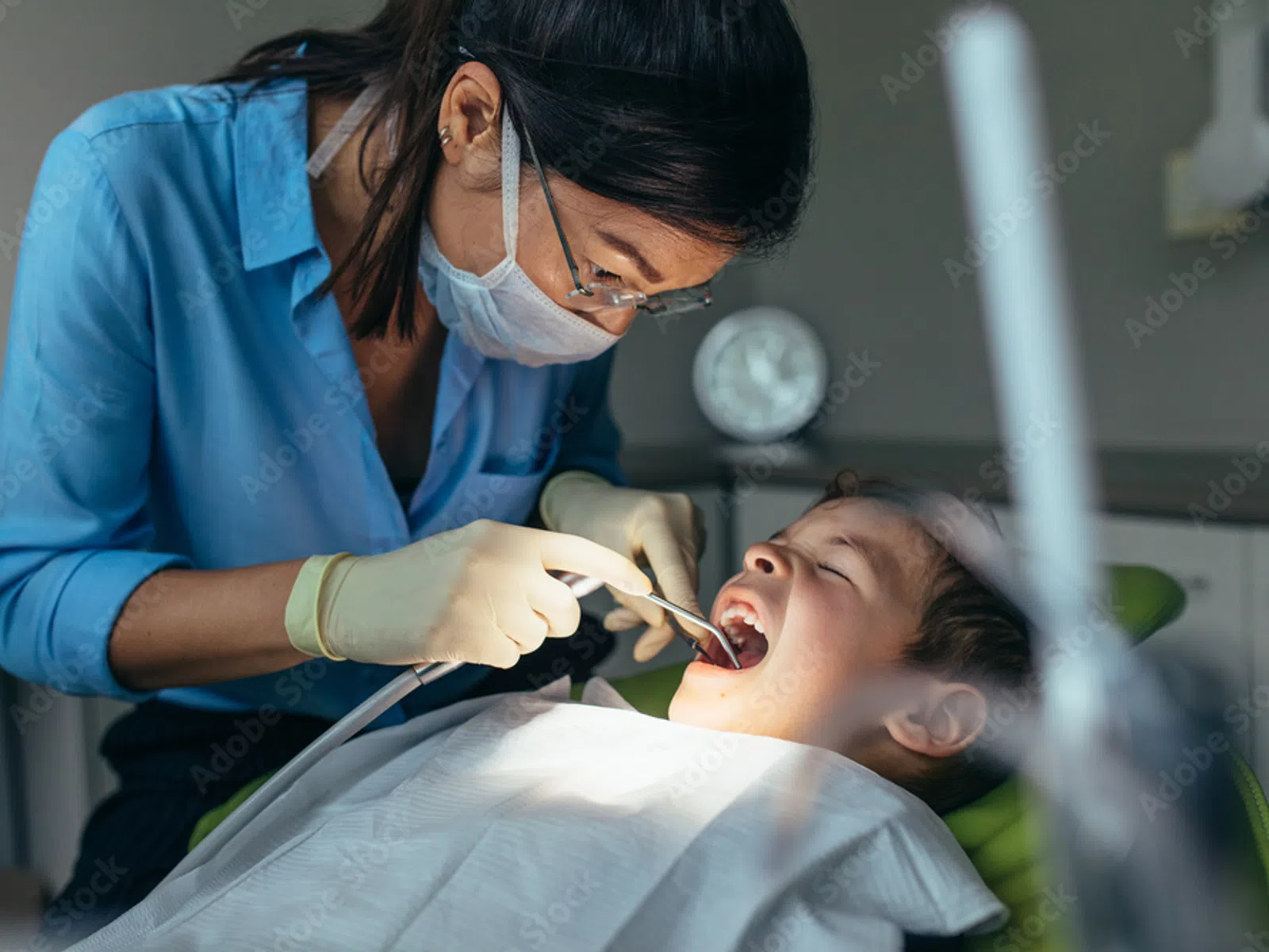 Dentist wearing mask examines young boy's teeth during dental checkup in clinic with focused lighting.