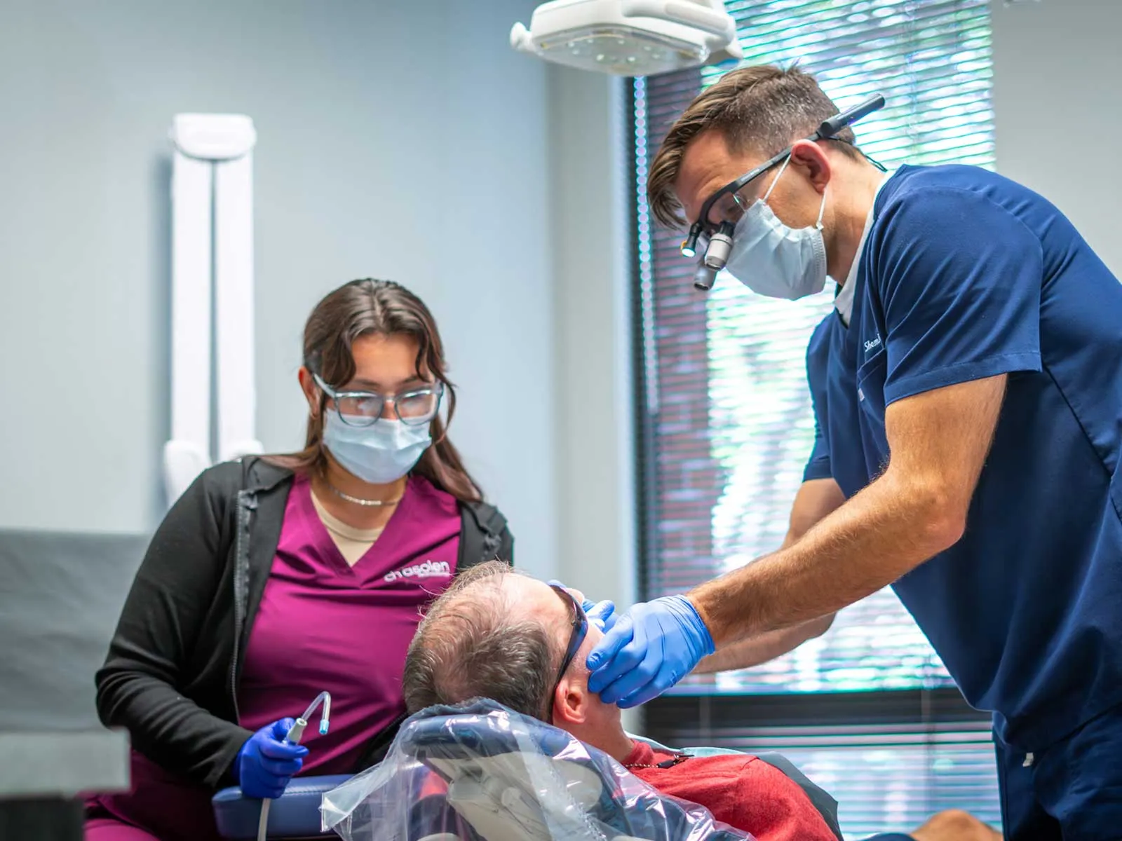 Dentist and assistant wearing masks and gloves treating a patient in a dental clinic with modern equipment.