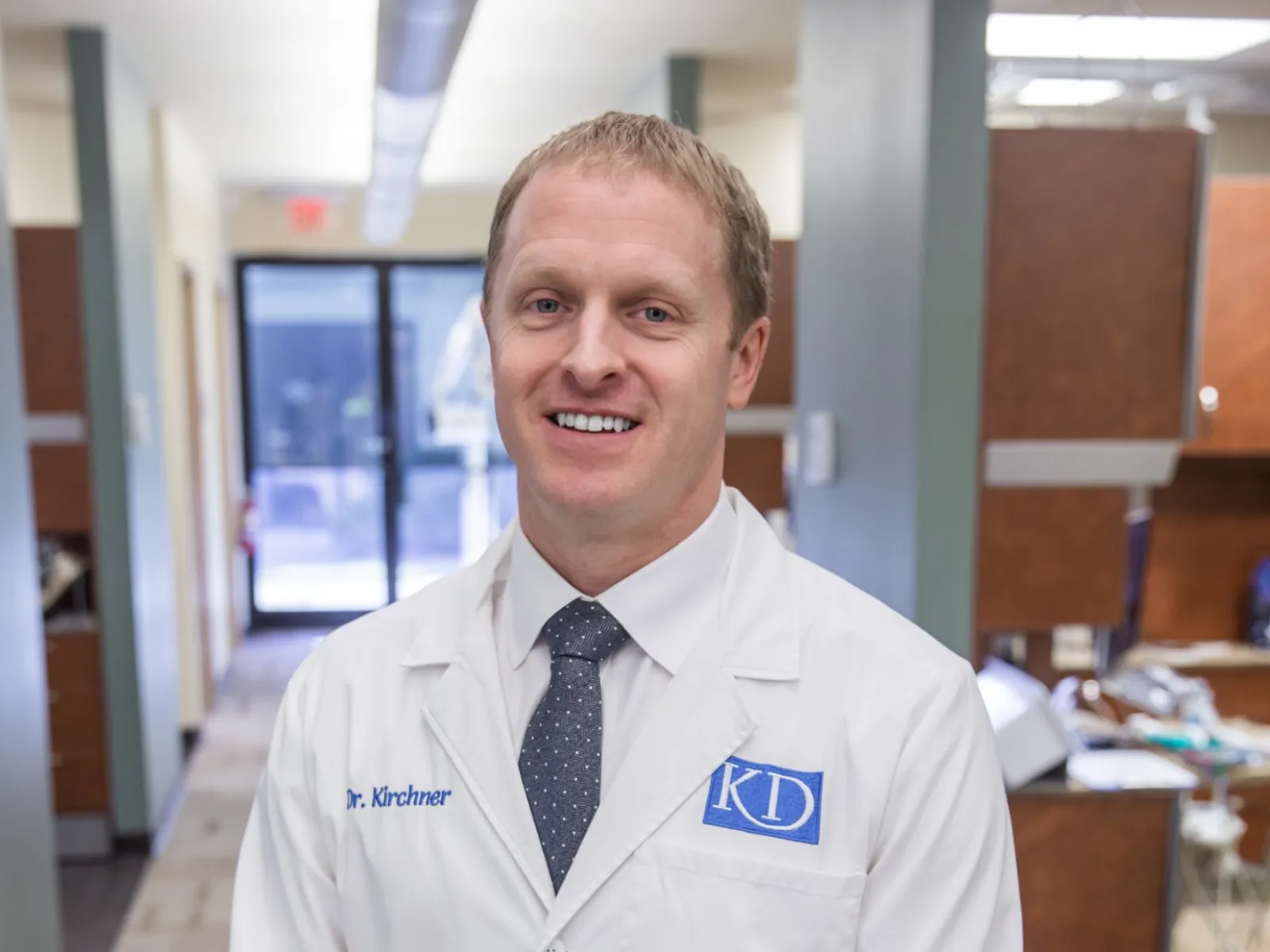 Smiling male doctor in white lab coat standing in a modern medical office hallway.