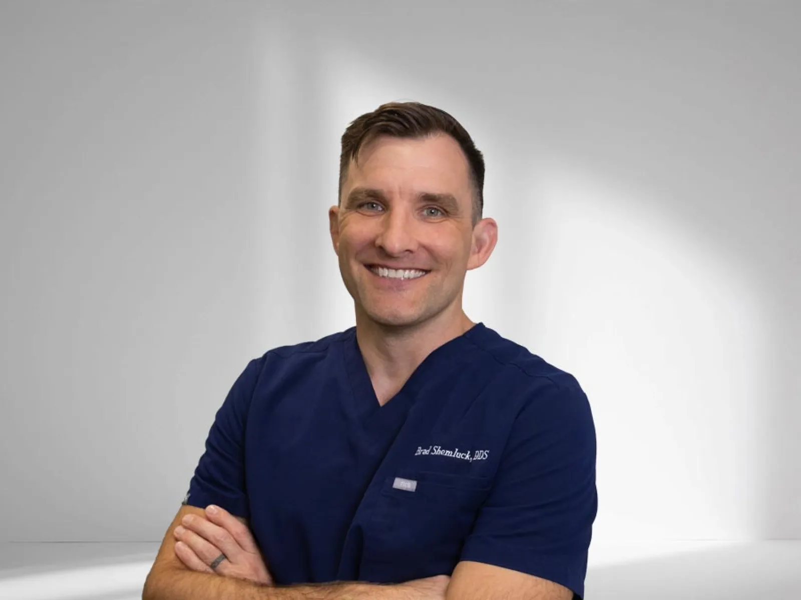 Smiling male dentist in navy blue scrubs with arms crossed against a plain light background.